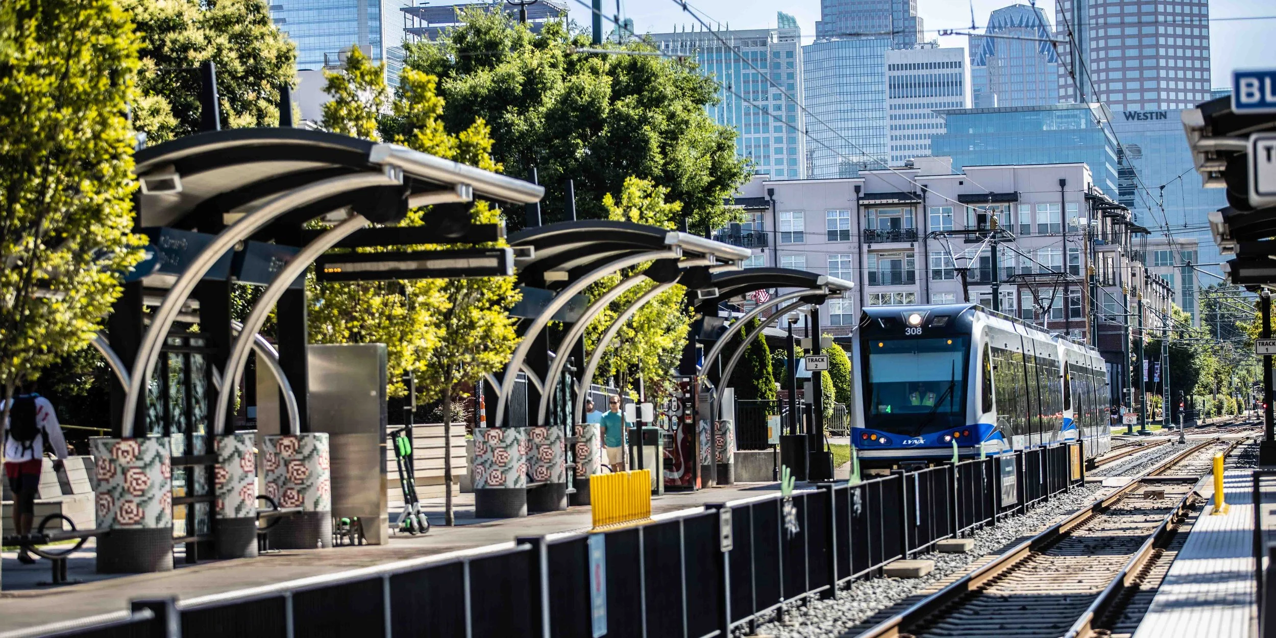 A city train station with a modern train arriving on the tracks, surrounded by green trees and tall skyscrapers in the background.