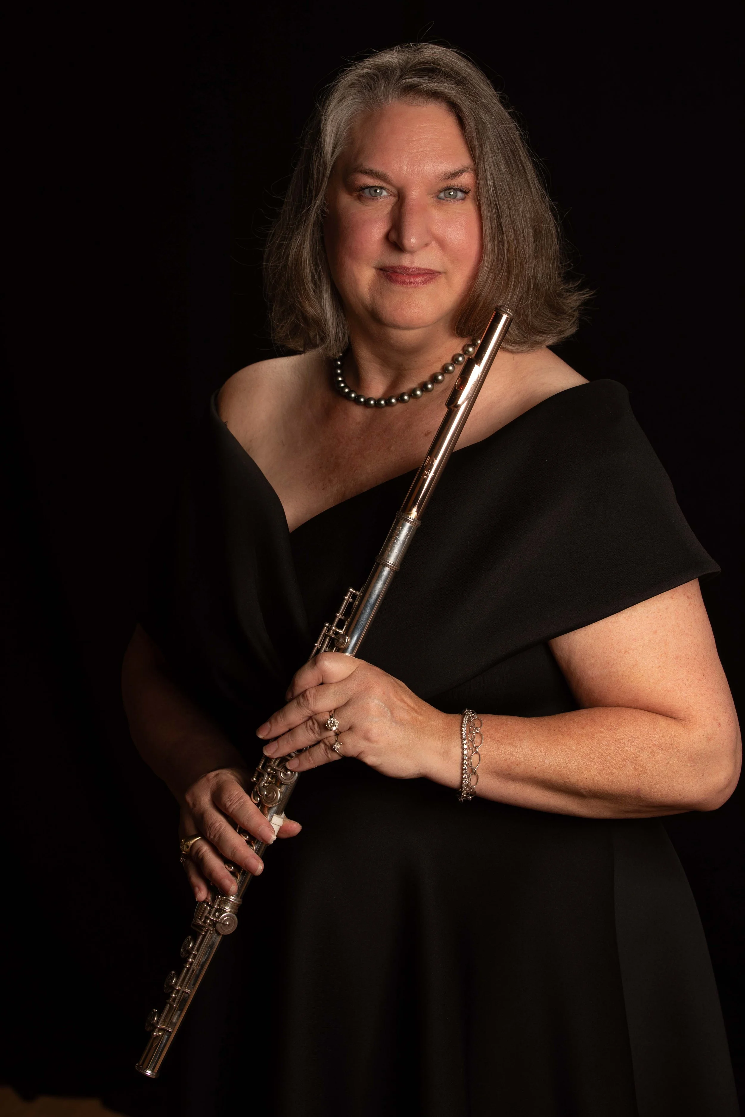 A middle-aged woman with shoulder-length gray hair and blue eyes is wearing a black off-shoulder dress, a pearl necklace, and bracelets, and is holding a silver flute against a black background.