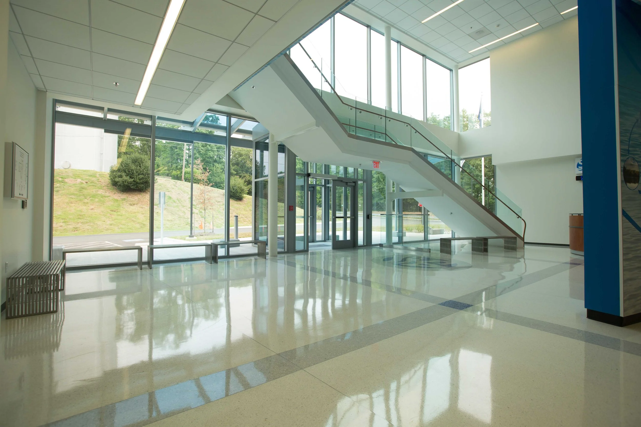 Empty modern lobby area with large glass windows, staircase, and seating benches, with outdoors visible through the windows.