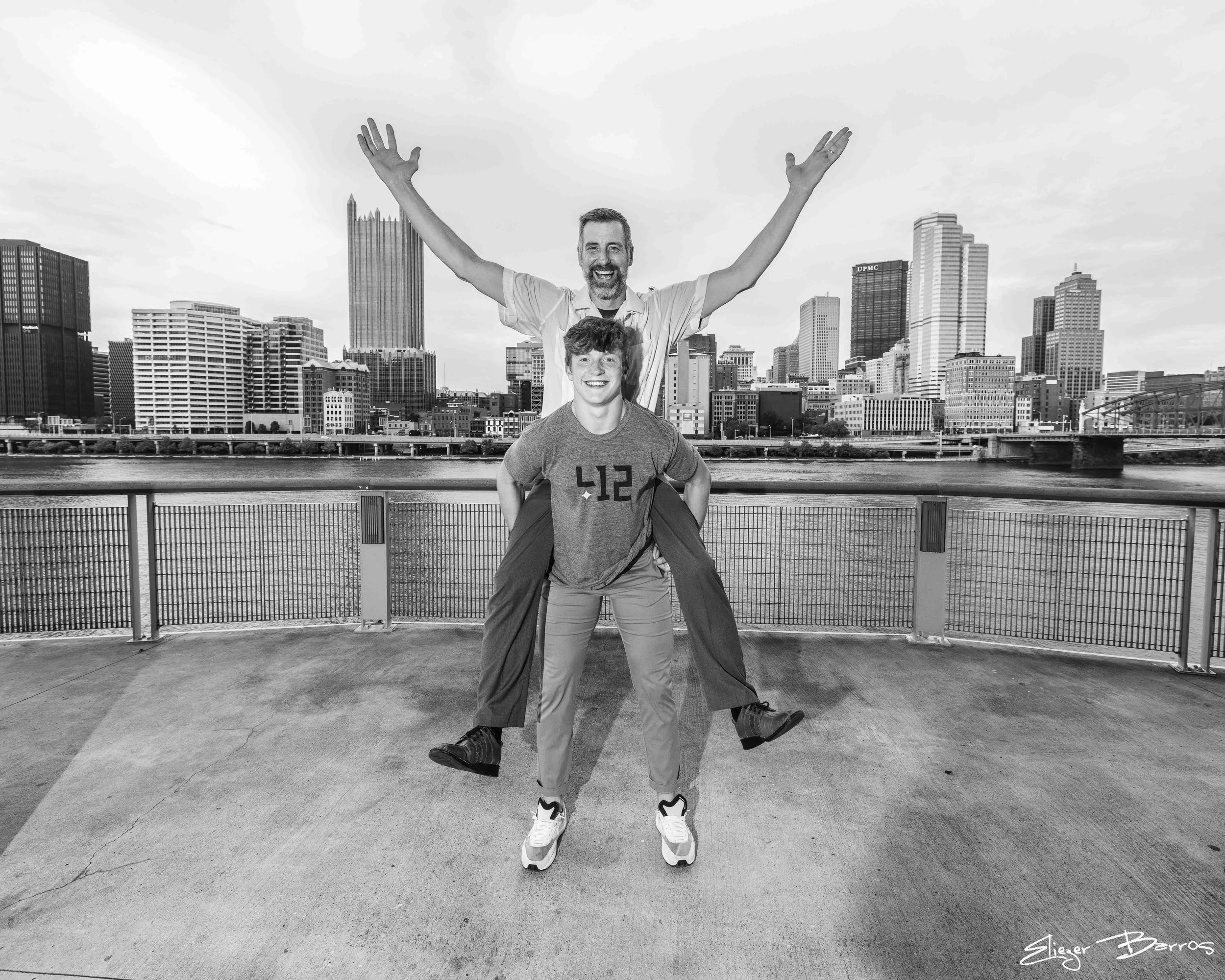 A man with a beard and a smile, lifting a young person wearing a hoodie, both standing on a bridge with a city skyline featuring tall buildings in the background.
