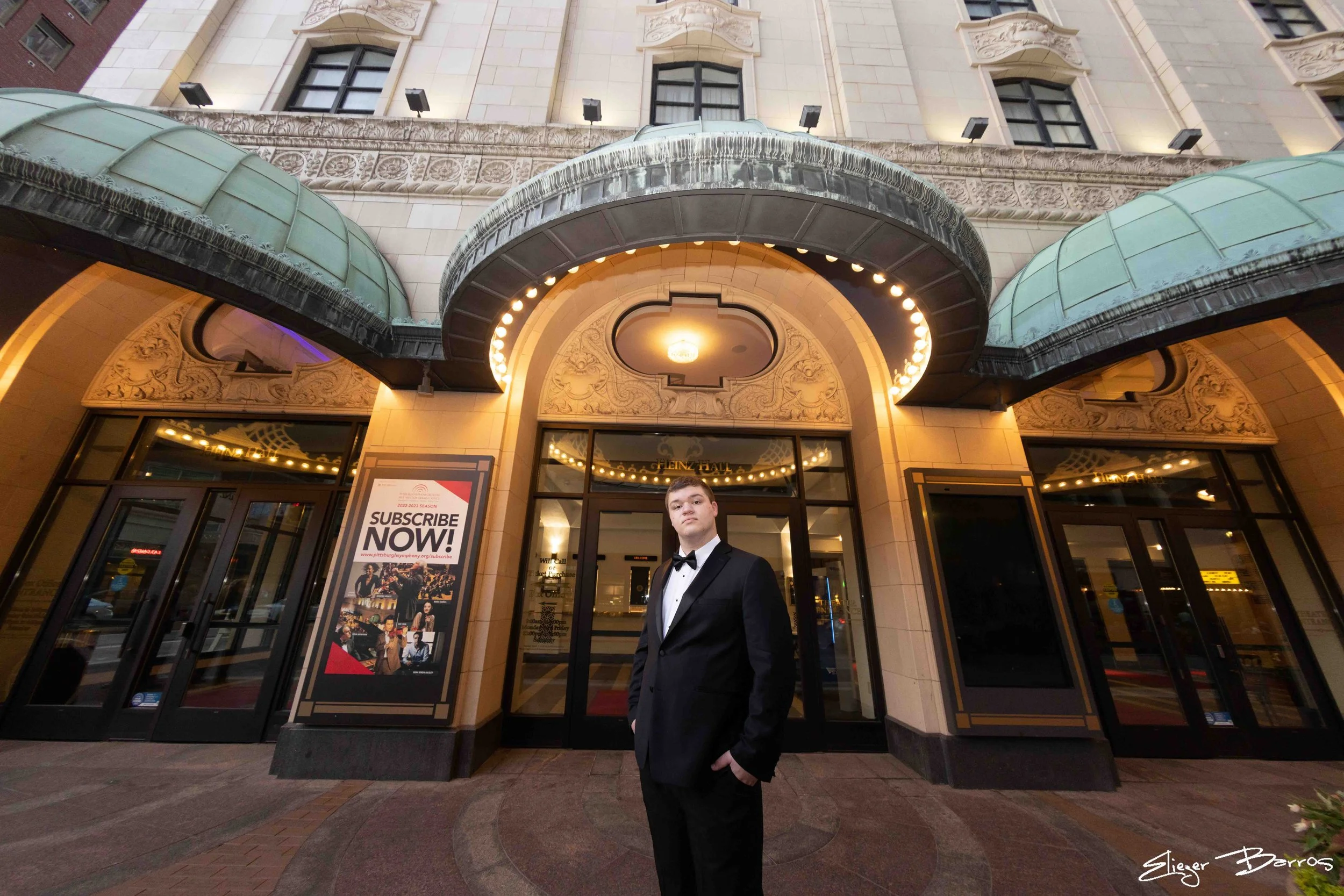 A young man in a tuxedo standing in front of the entrance to a theater with ornate architectural details, large glass doors, and a promotional poster for a show called Pittburgh Symphony.