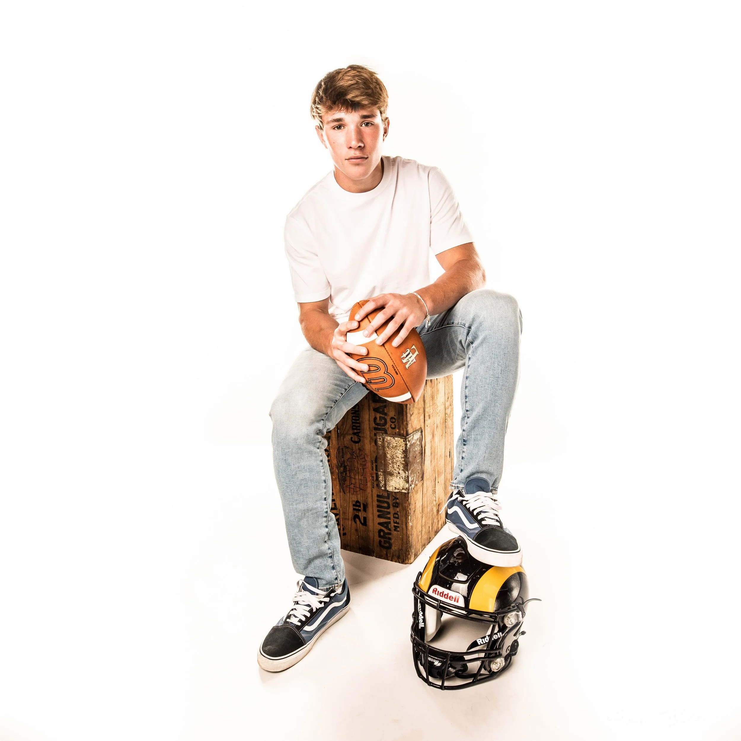 Young man sitting on a wooden crate, holding a football, with a football helmet and a miniature football helmet on the floor in front of him, against a white background.