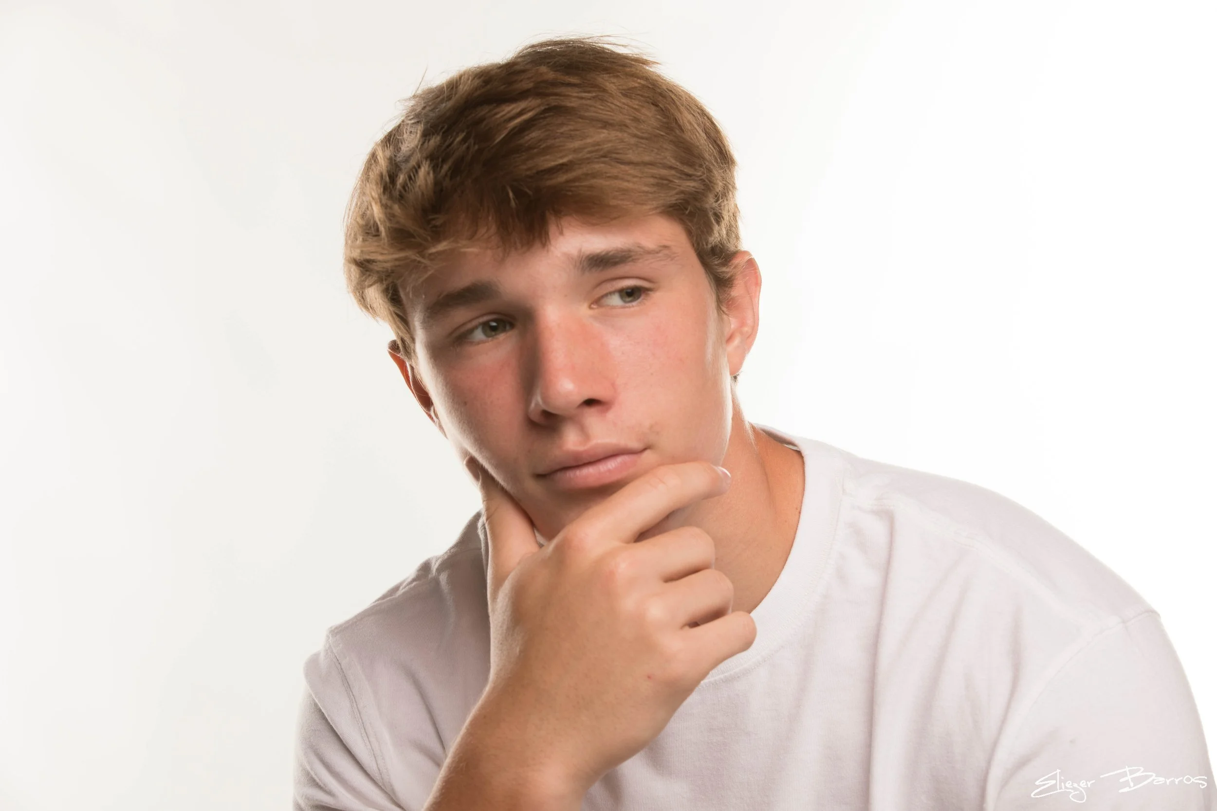 Young man with light brown hair and blue eyes, wearing a white shirt, resting his chin on his hand and looking slightly to the side, against a plain white background.