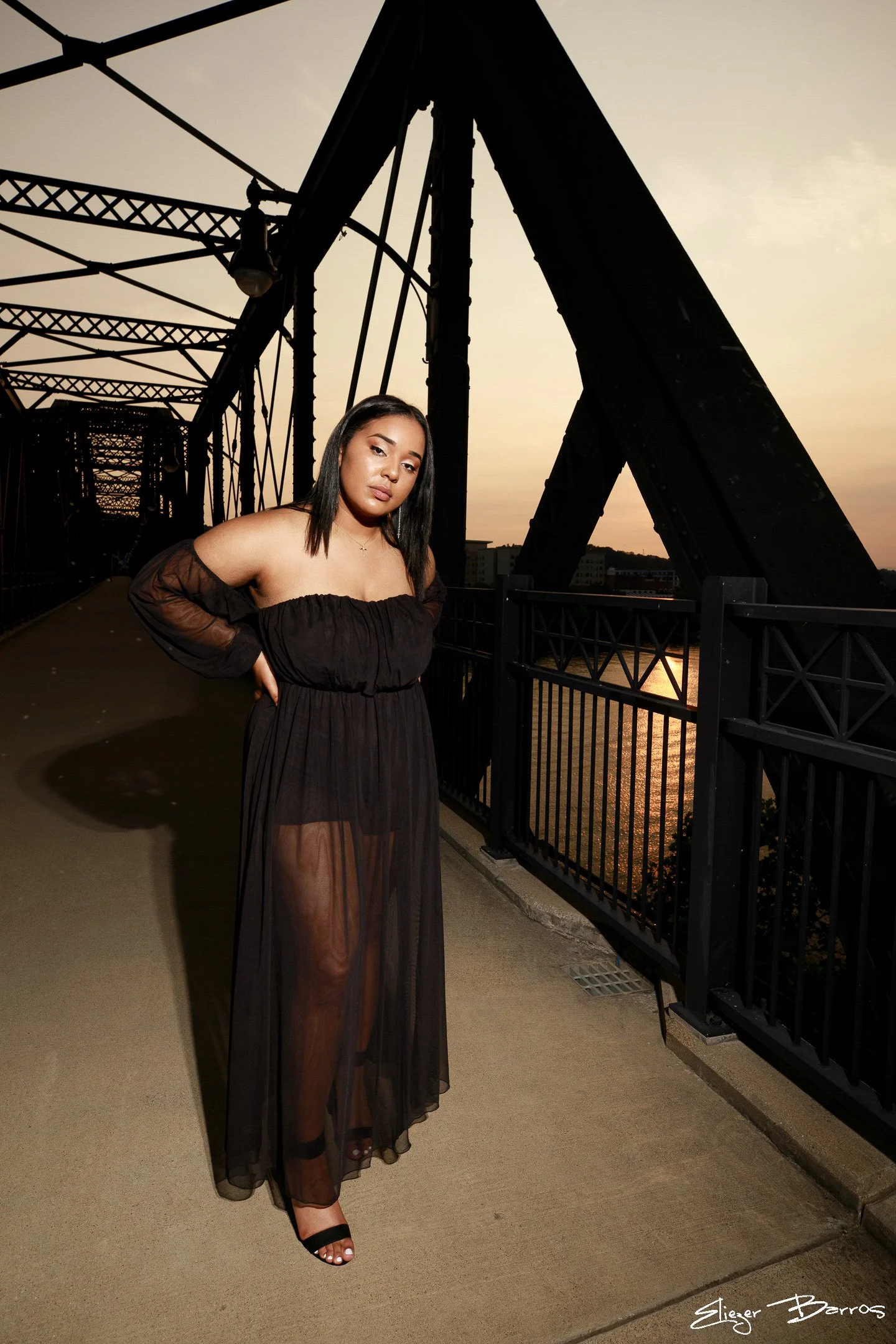 A woman wearing a black off-the-shoulder dress with a sheer overlay stands on a bridge during sunset. She has shoulder-length dark hair and is wearing black high-heeled sandals. The bridge has metal framework visible in the background, and the sky is