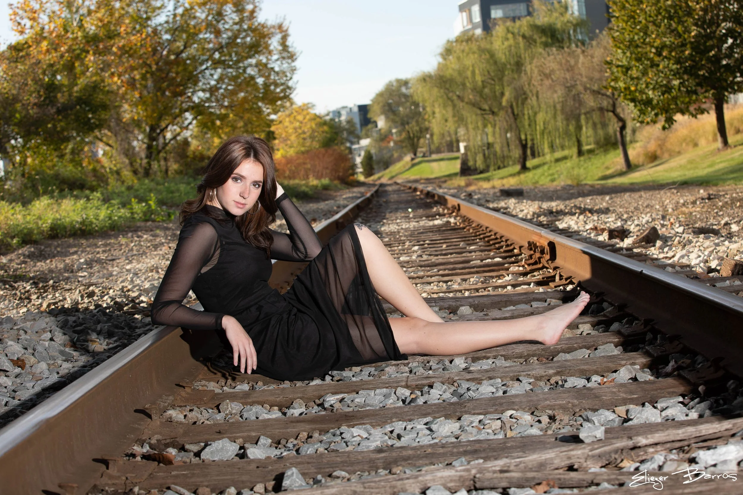 A woman with brown hair in a black sheer dress sits on railroad tracks in a park with trees and buildings in the background.