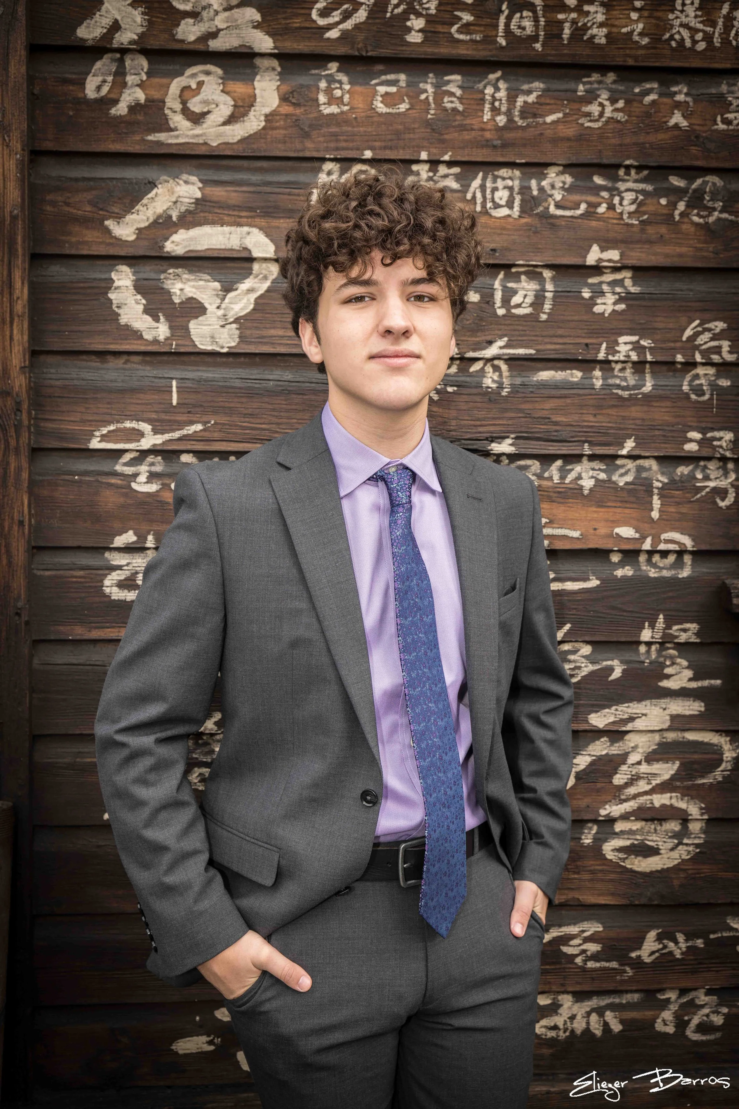 A young man with curly hair wearing a grey suit, light purple shirt, and a blue patterned tie, standing with one hand in his pocket in front of a wooden wall with white Chinese characters painted on it.