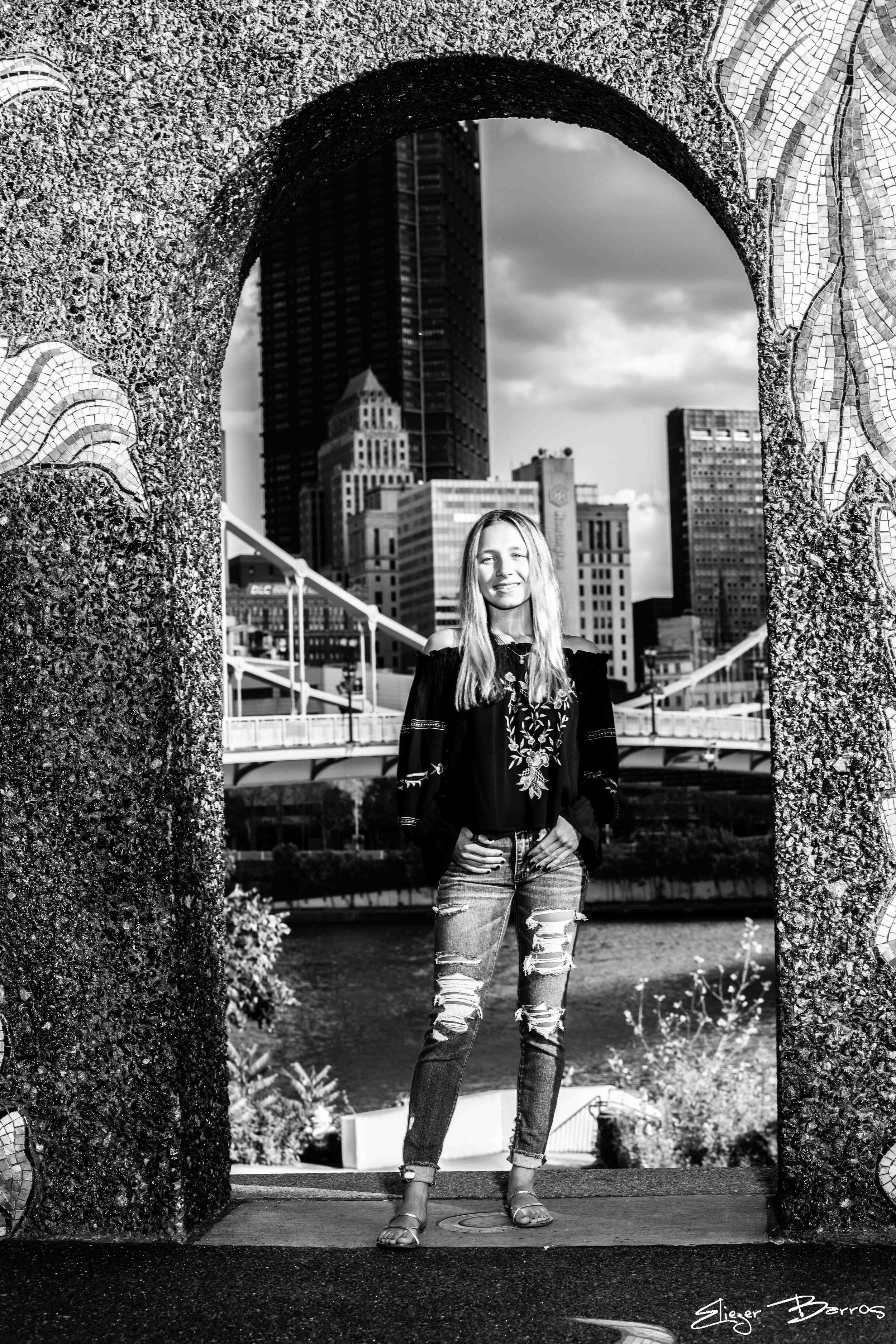 Black and white photo of a young woman standing between two textured stone columns with a city skyline, including skyscrapers, in the background. She is smiling, wearing ripped jeans, a floral top, and sandals.