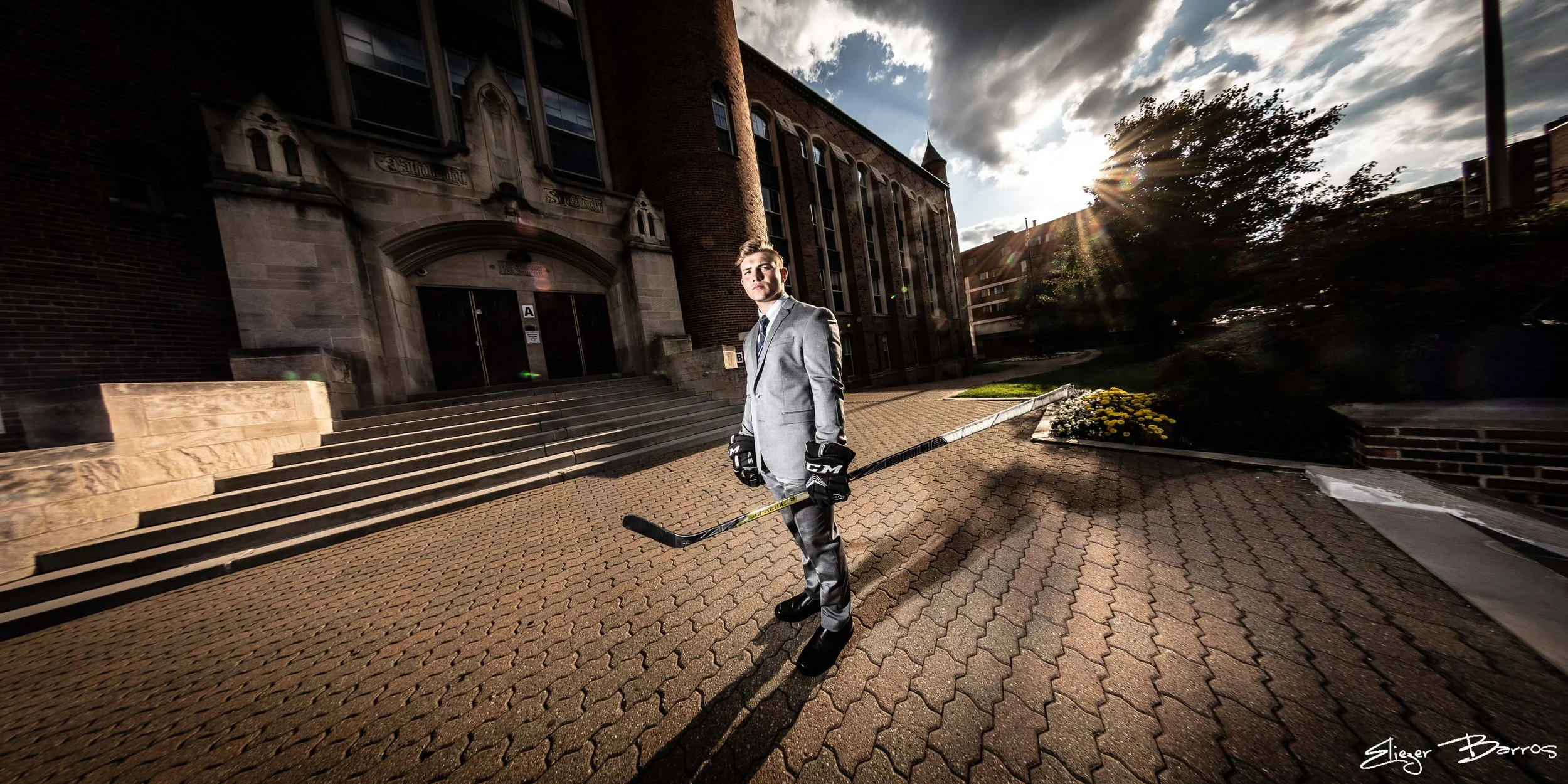 A man in a gray suit and ice hockey gloves stands on a brick sidewalk holding a hockey stick in front of a historic building with stairs, with the sun setting behind clouds in the sky.