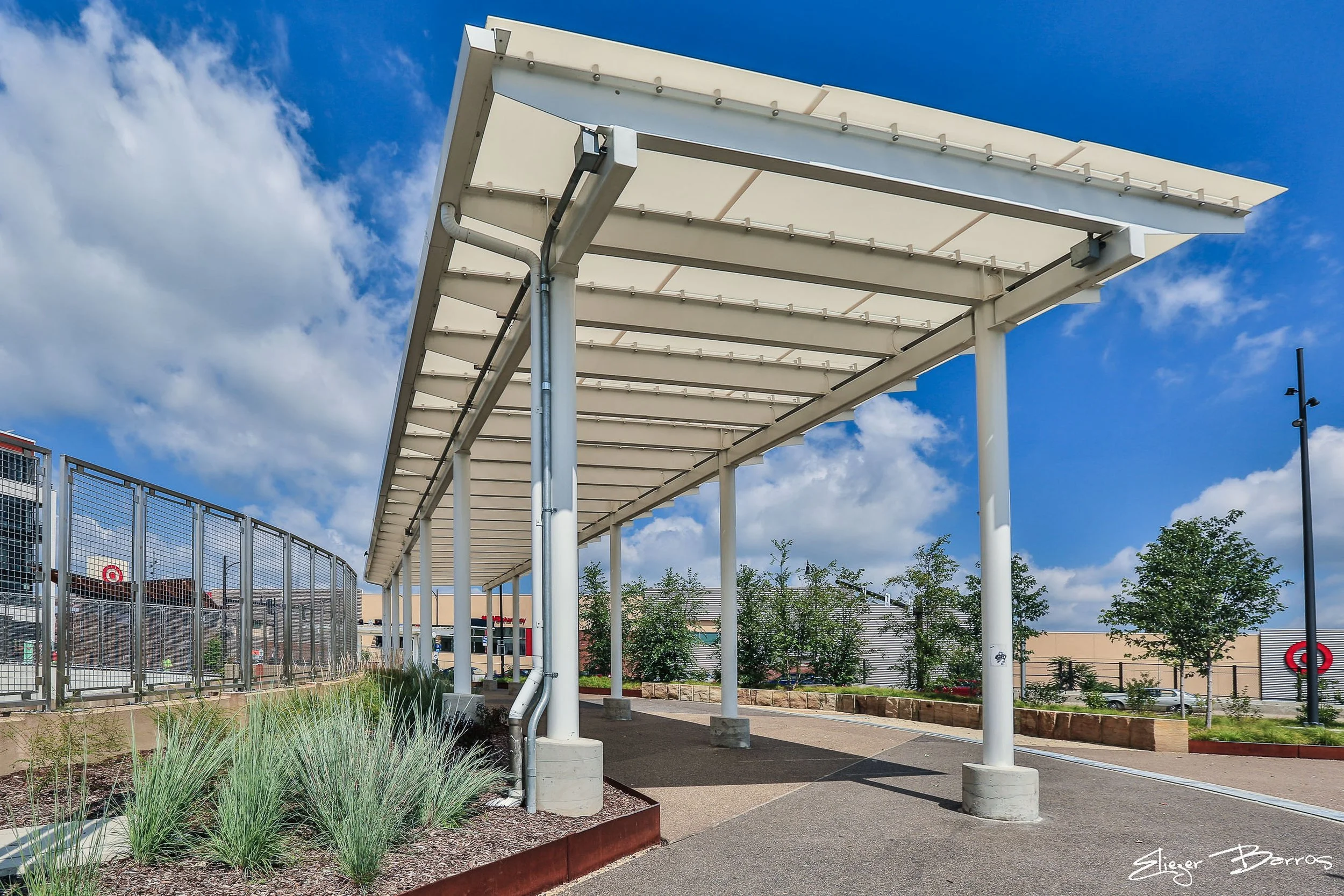 A modern outdoor canopy with white metal supports and a translucent roof, located in a shopping or commercial area. Behind the canopy are retail stores including Target, with fencing along the sidewalk and some greenery in the foreground. The sky is 