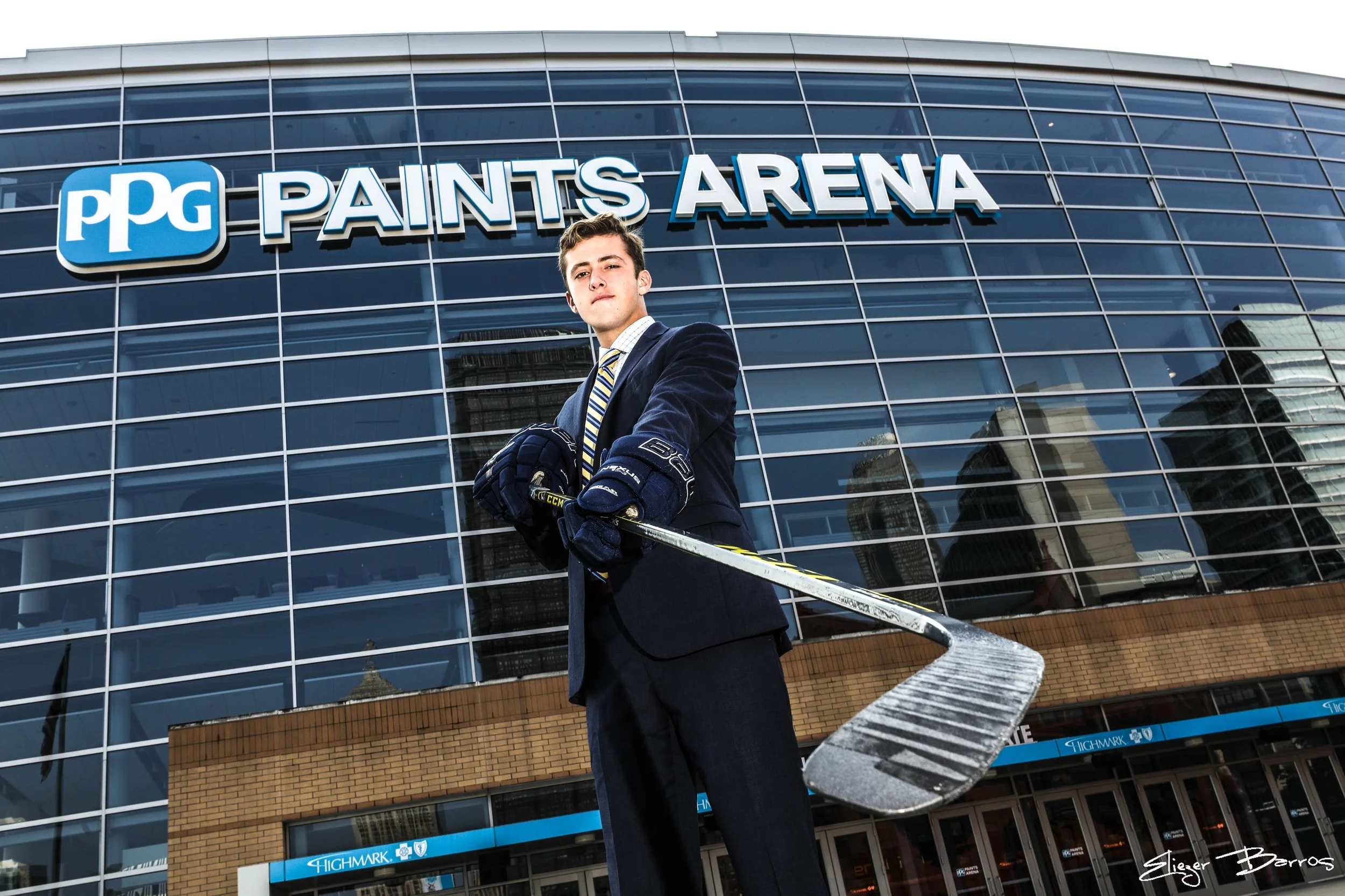 A young man in a suit and hockey gloves standing in front of the PPG Paints Arena, a glass building with blue signage.