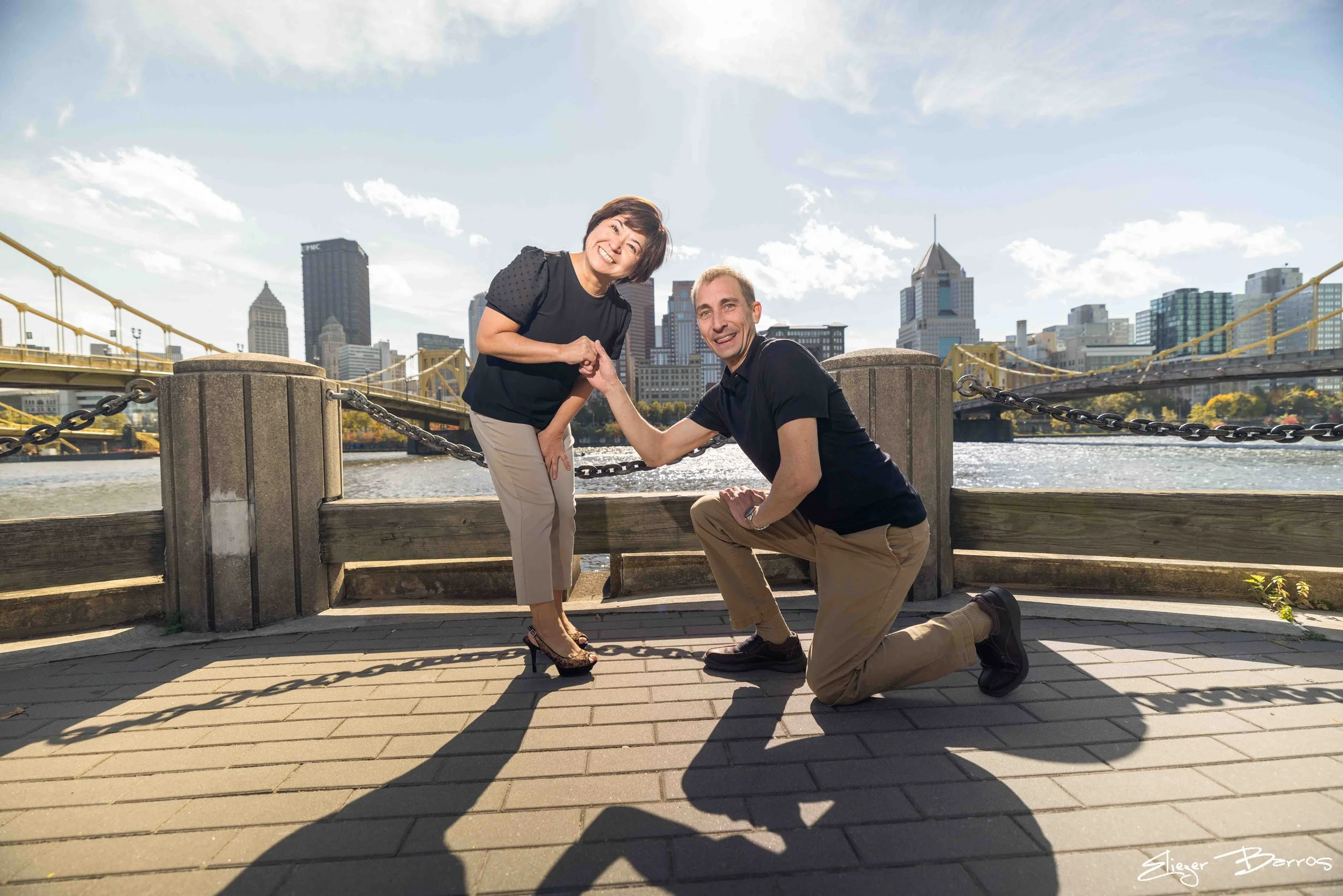 A man kneeling on one knee, holding hands with a smiling woman standing beside him near a river, with a city skyline and bridge in the background on a sunny day.