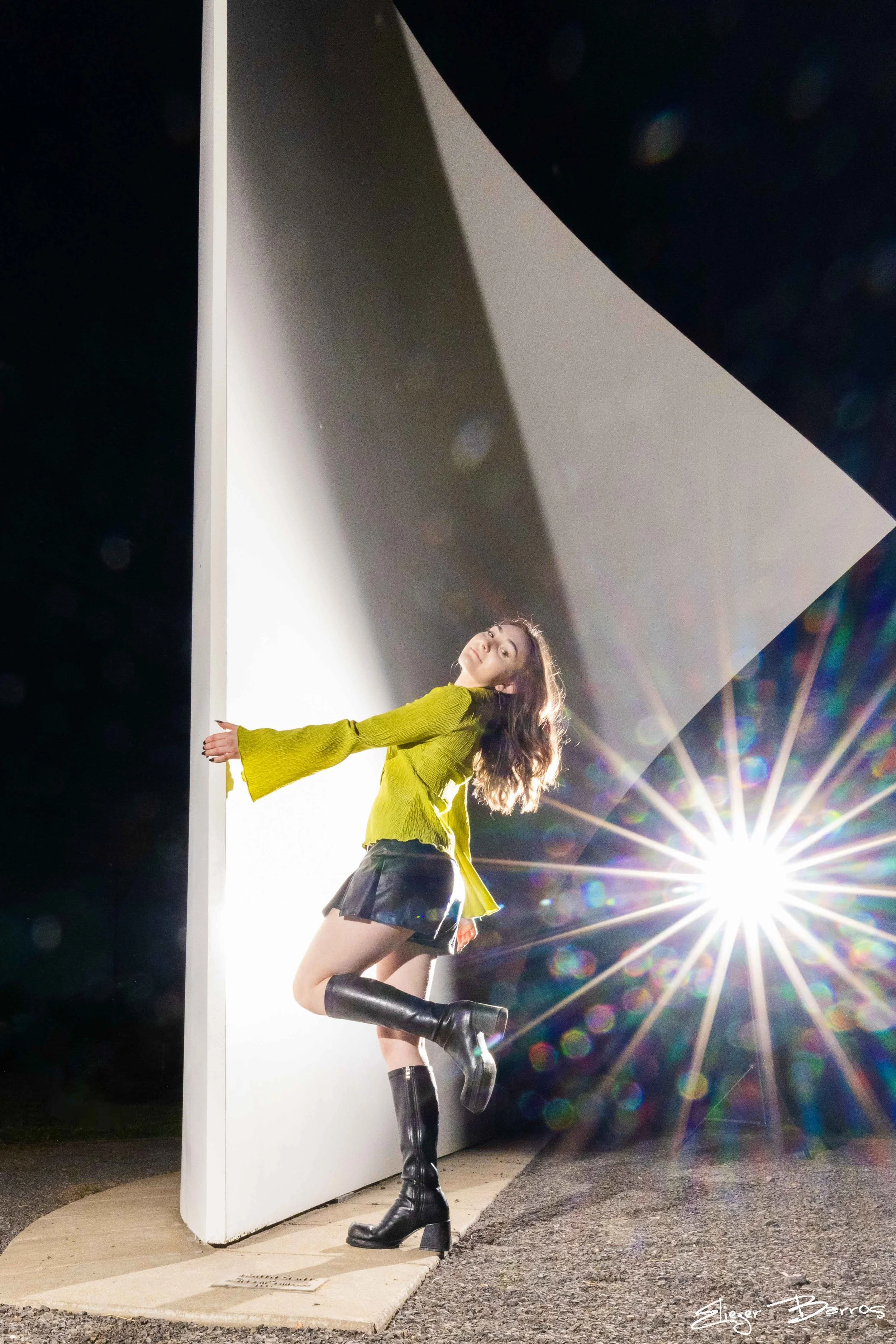 A young woman with long, wavy hair and a confident expression poses against a white panel outdoors at night. She is wearing a bright yellow top with flared sleeves, a black mini skirt, and knee-high black leather boots. The sun or a strong light sour