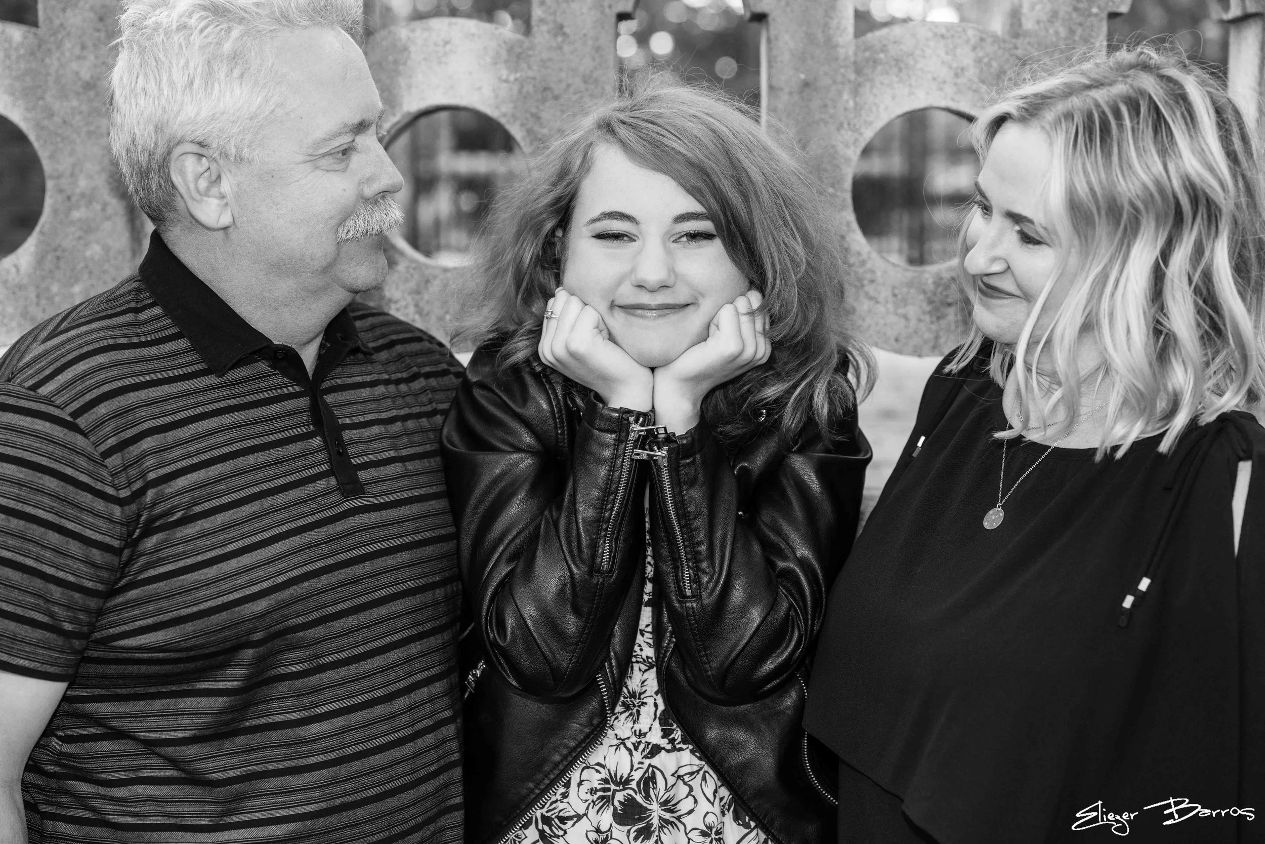 Three people standing close together outdoors in front of an animal-shaped concrete sculpture. The person in the middle, a young woman, is smiling with her chin resting on her hands, wearing a leather jacket and floral dress. The person on the left i