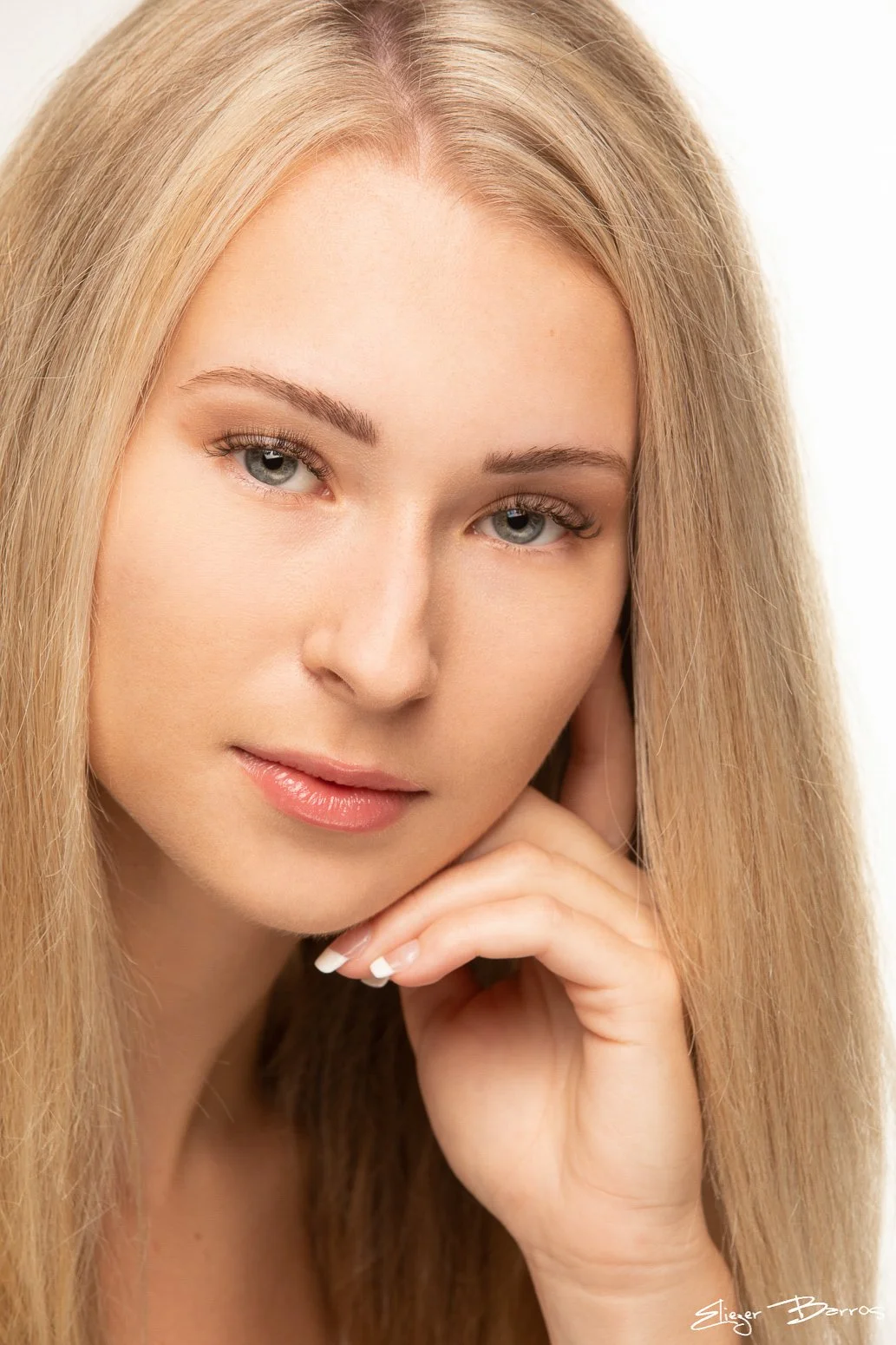 Close-up portrait of a woman with long blonde hair and light-colored eyes, resting her chin on her hand, against a light background.