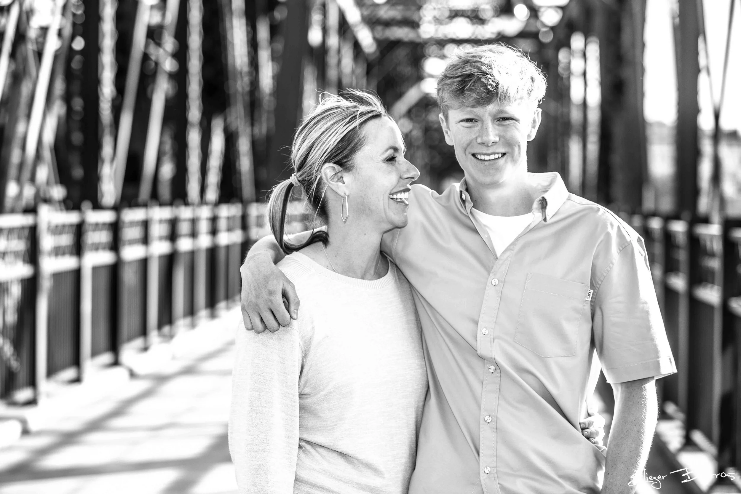 A woman and a young man are smiling and embracing on a bridge with metal railings, outdoors on a sunny day.