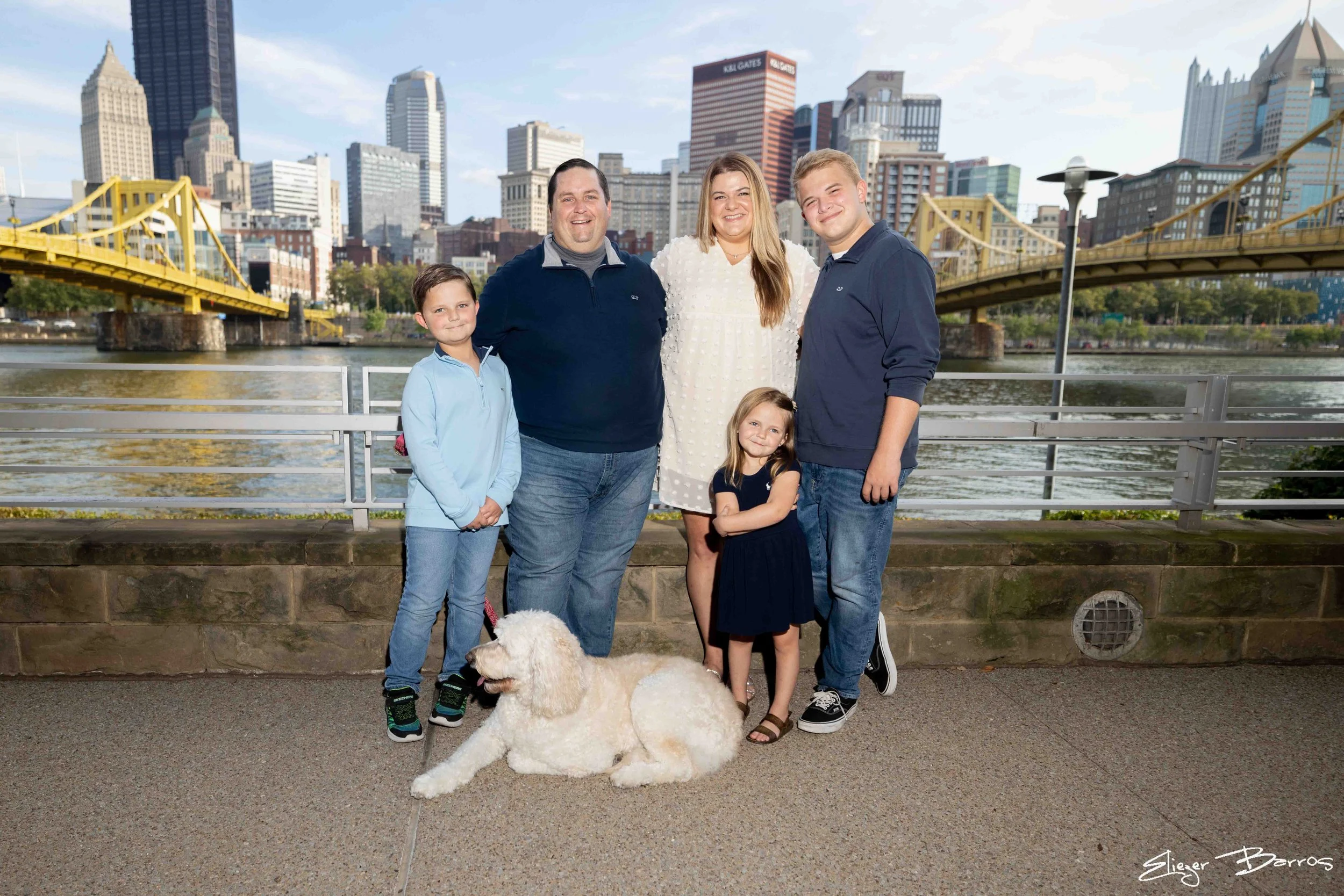 Family of six with two young girls and their three adult men standing by a river with a city skyline in the background, and a large white dog laying on the ground in front.