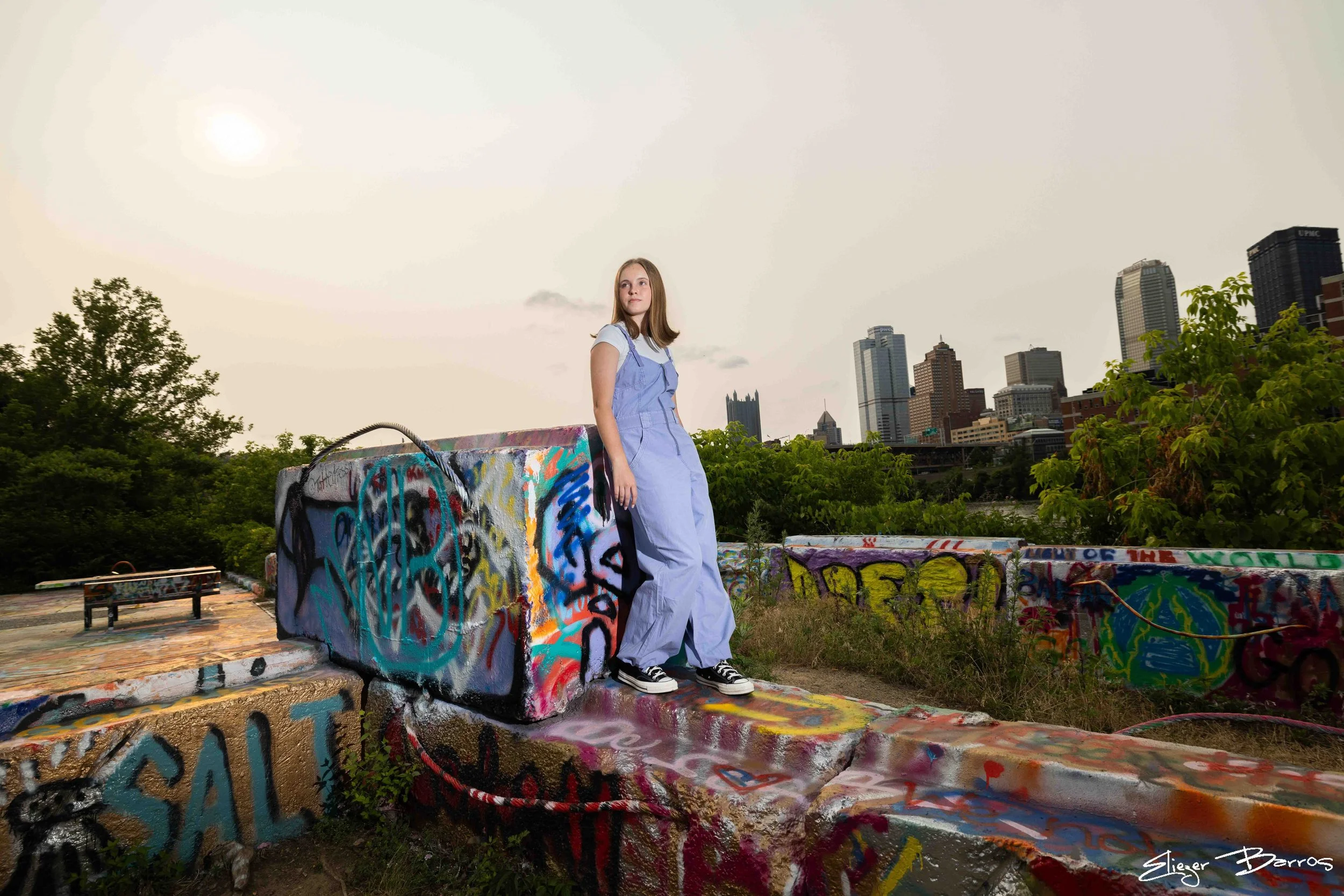 Senior Pictures Pittsburgh at Highline & Color Park. Young woman in a light blue dress posing on colorful graffiti-covered structures with Pittsburgh skyline and river views for artistic outdoor senior portraits.