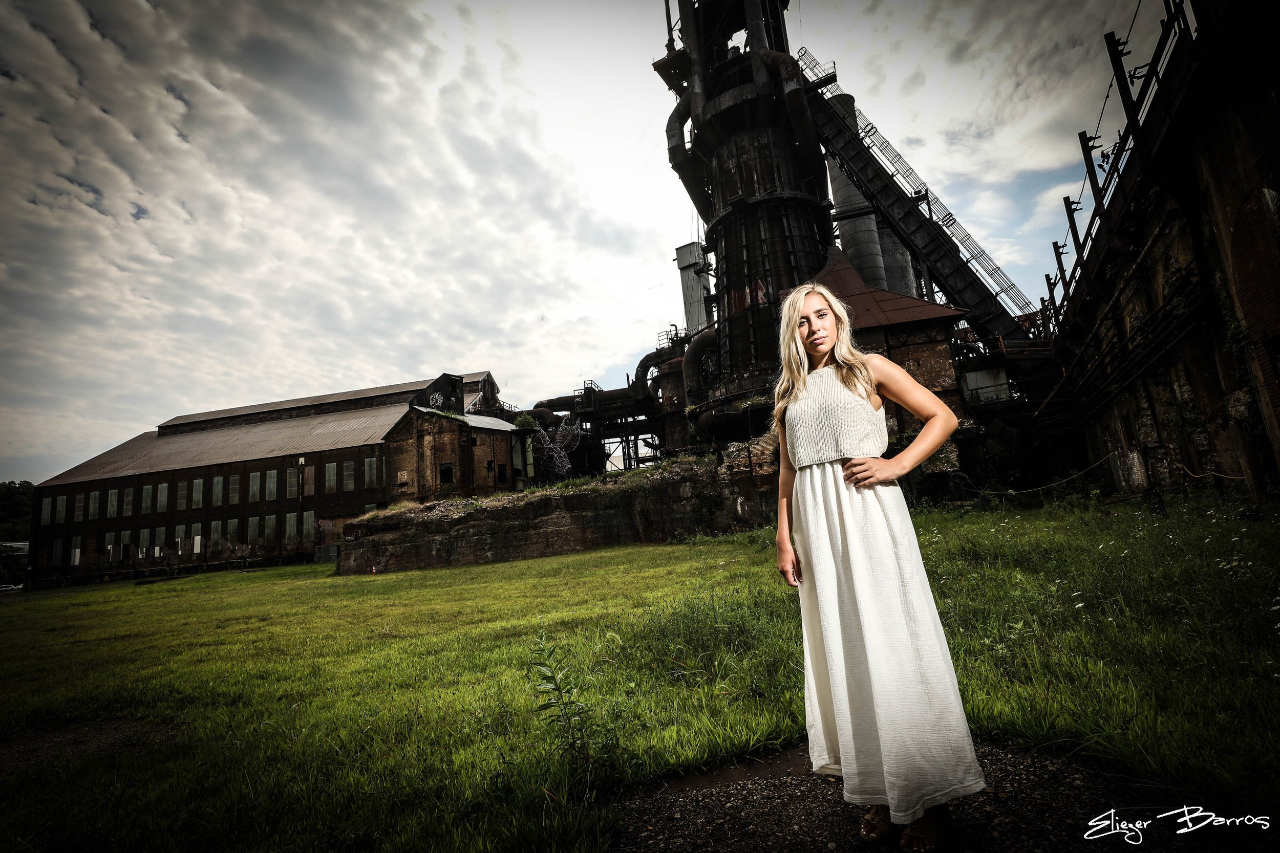A woman in a long white dress standing outdoors on green grass in front of an industrial factory with tall structures and pipes, under a cloudy sky.