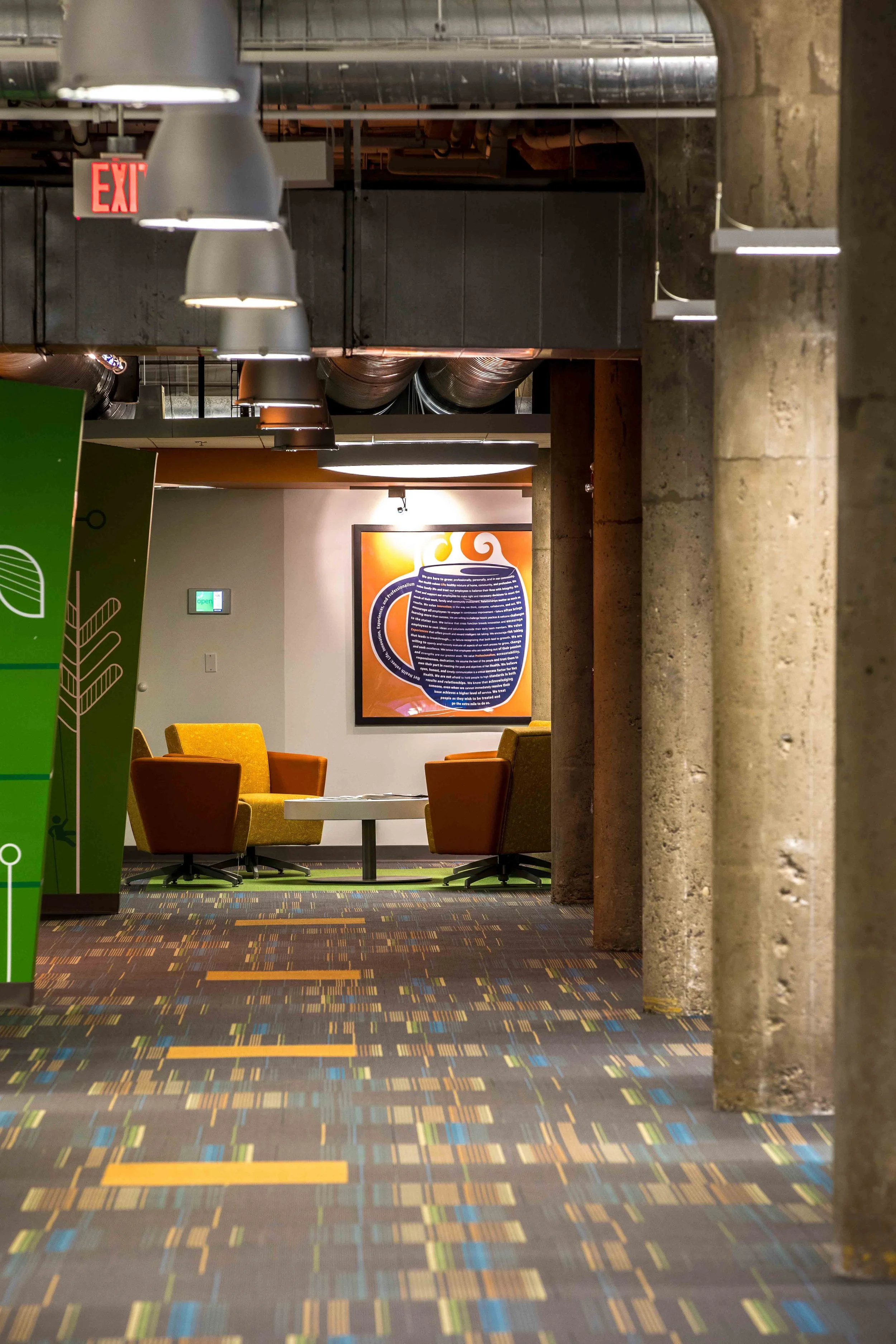 A modern office lounge area with colorful chairs, a framed abstract artwork, and patterned carpet. There are exposed ducts and industrial style lighting.