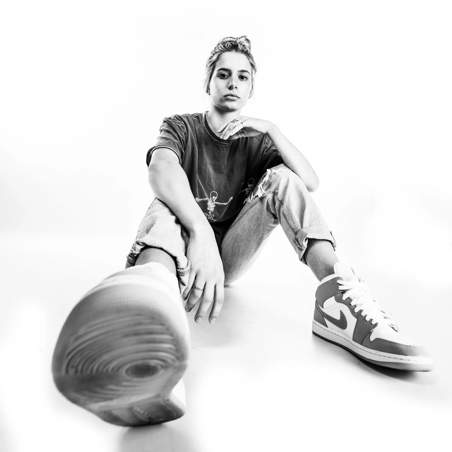 Black and white photo of a young woman sitting on the floor, wearing a t-shirt and jeans, with a sneaker in the foreground and her other foot visible. The photo appears to be taken from a low angle.