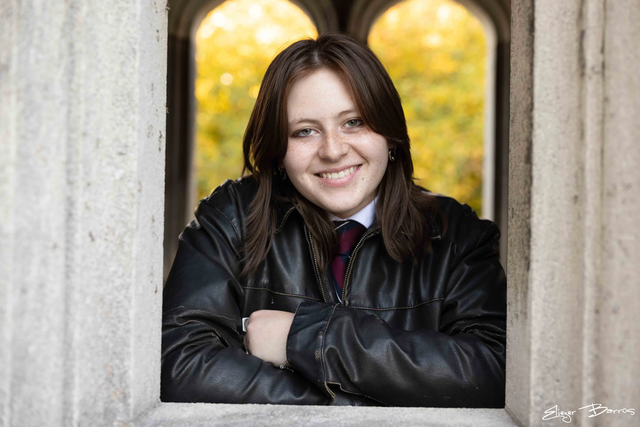 A young woman with brown hair and blue eyes smiling at the camera, leaning on her arms, wearing a leather jacket, with an autumn background visible through a stone window frame.