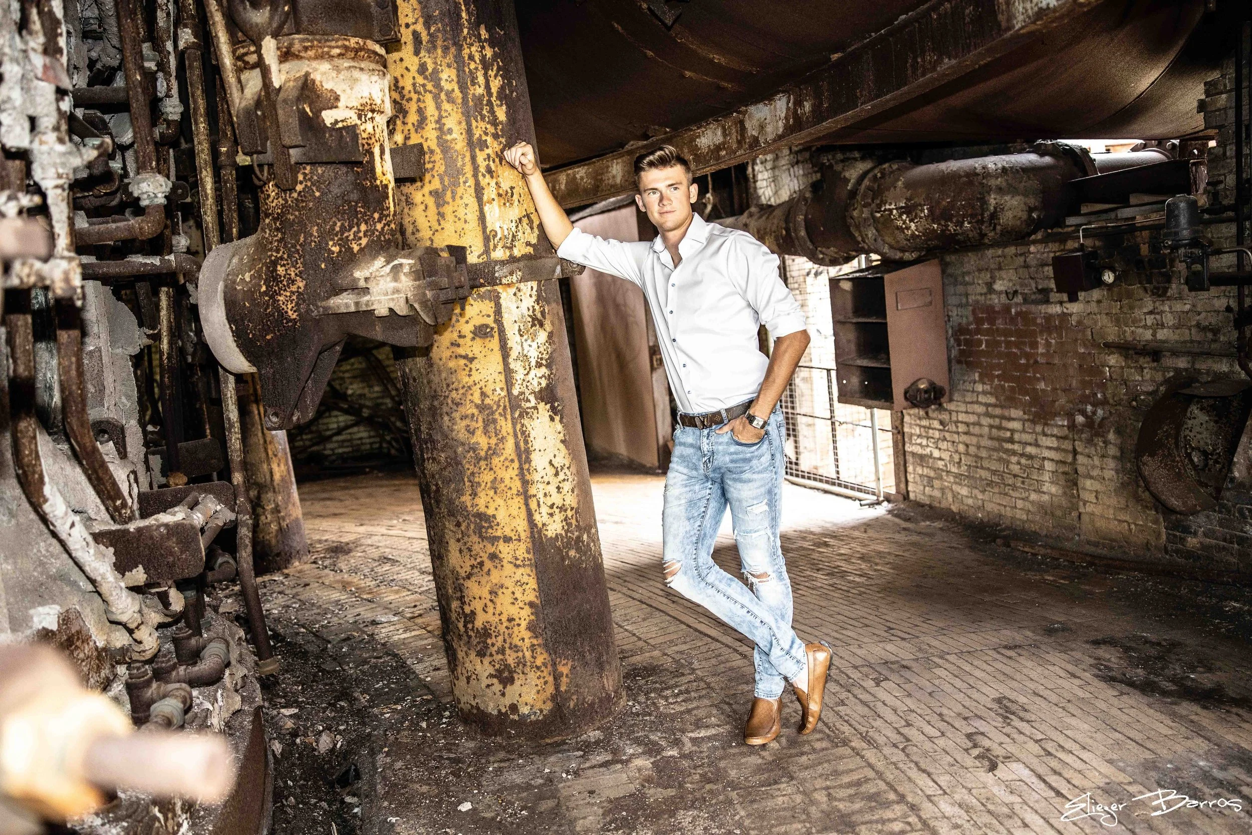 A young man in a white shirt and ripped jeans is standing inside a large industrial space with rusty machinery and pipes. He is resting his arm on a yellow metal component and looking confidently at camera. The environment is gritty with exposed bric