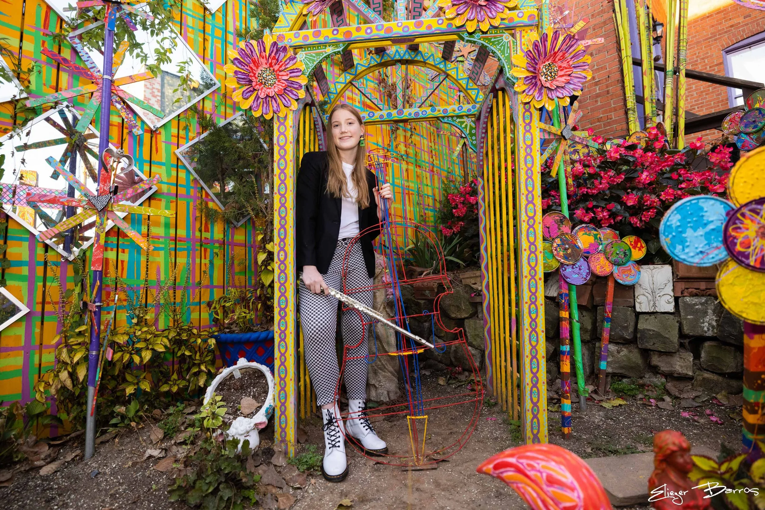 A young woman with long hair smiling, wearing a black blazer, white shirt, checkered pants, and white boots with black laces, stands in a colorful outdoor art installation with painted wooden structures, flowers, and mirrors.