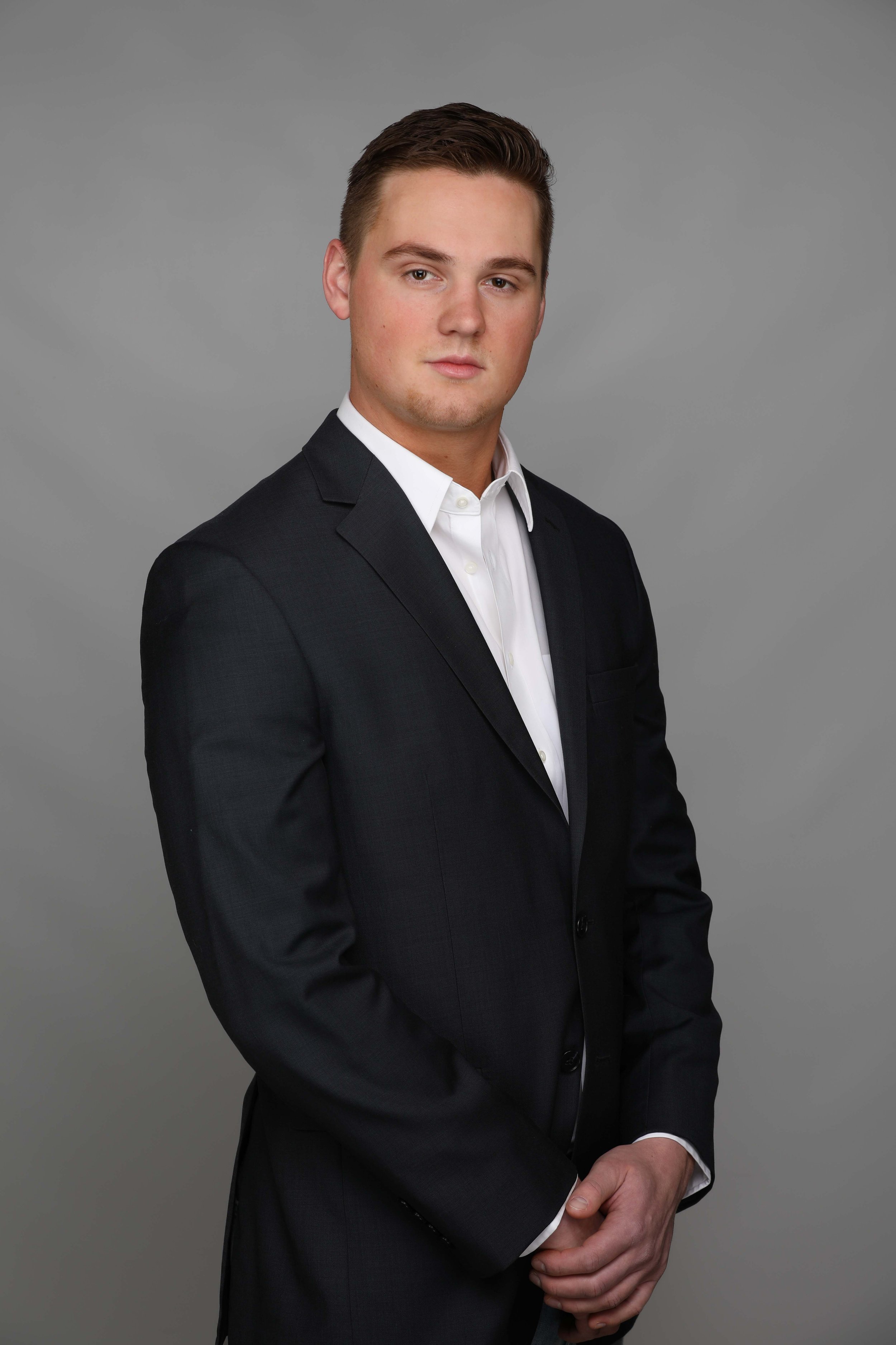 A young man with brown hair in a slicked-back style, wearing a black tuxedo jacket over a white dress shirt, standing against a gray background.