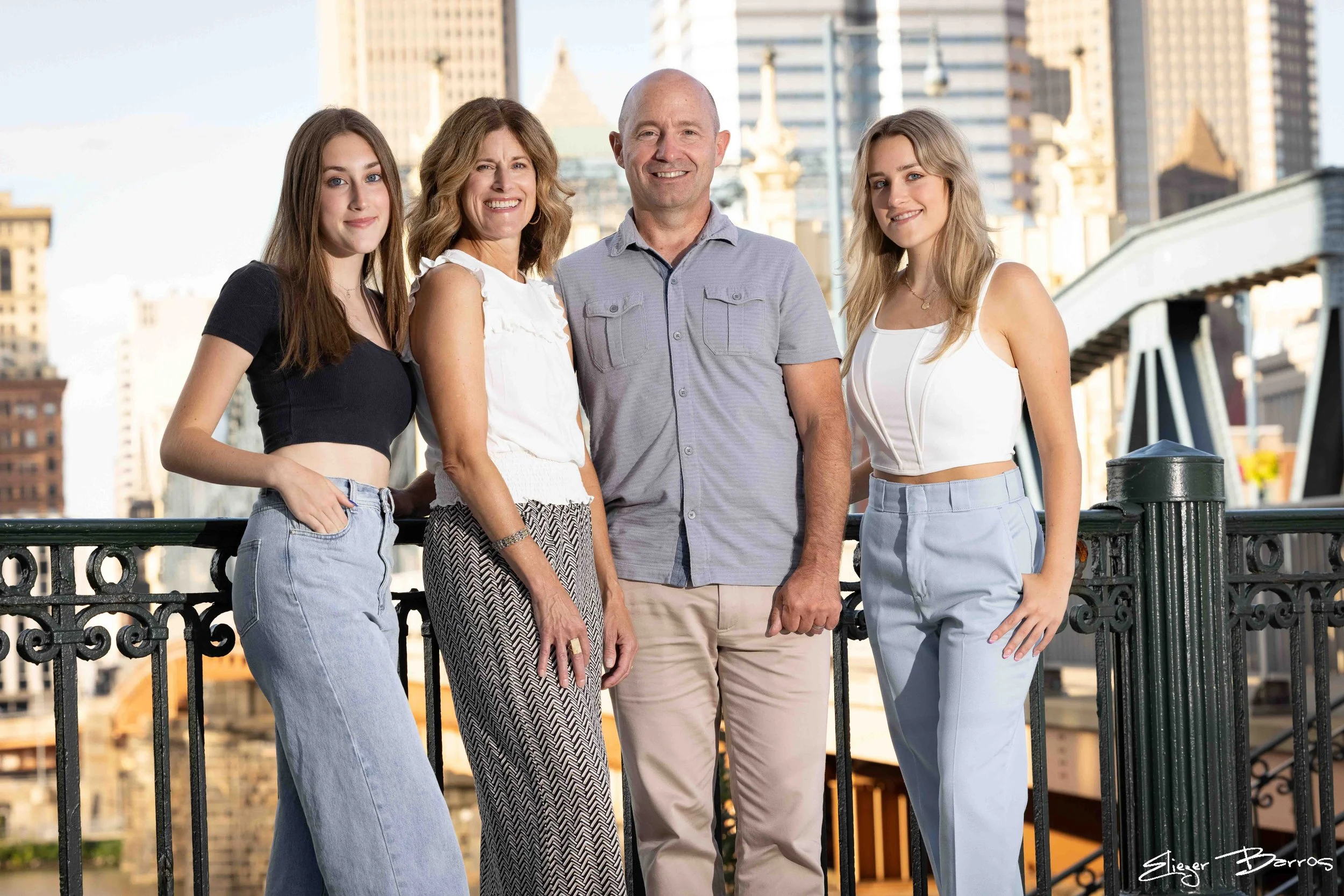 Four people standing together on a city balcony, with tall buildings in the background. They are smiling and facing the camera, dressed casually.