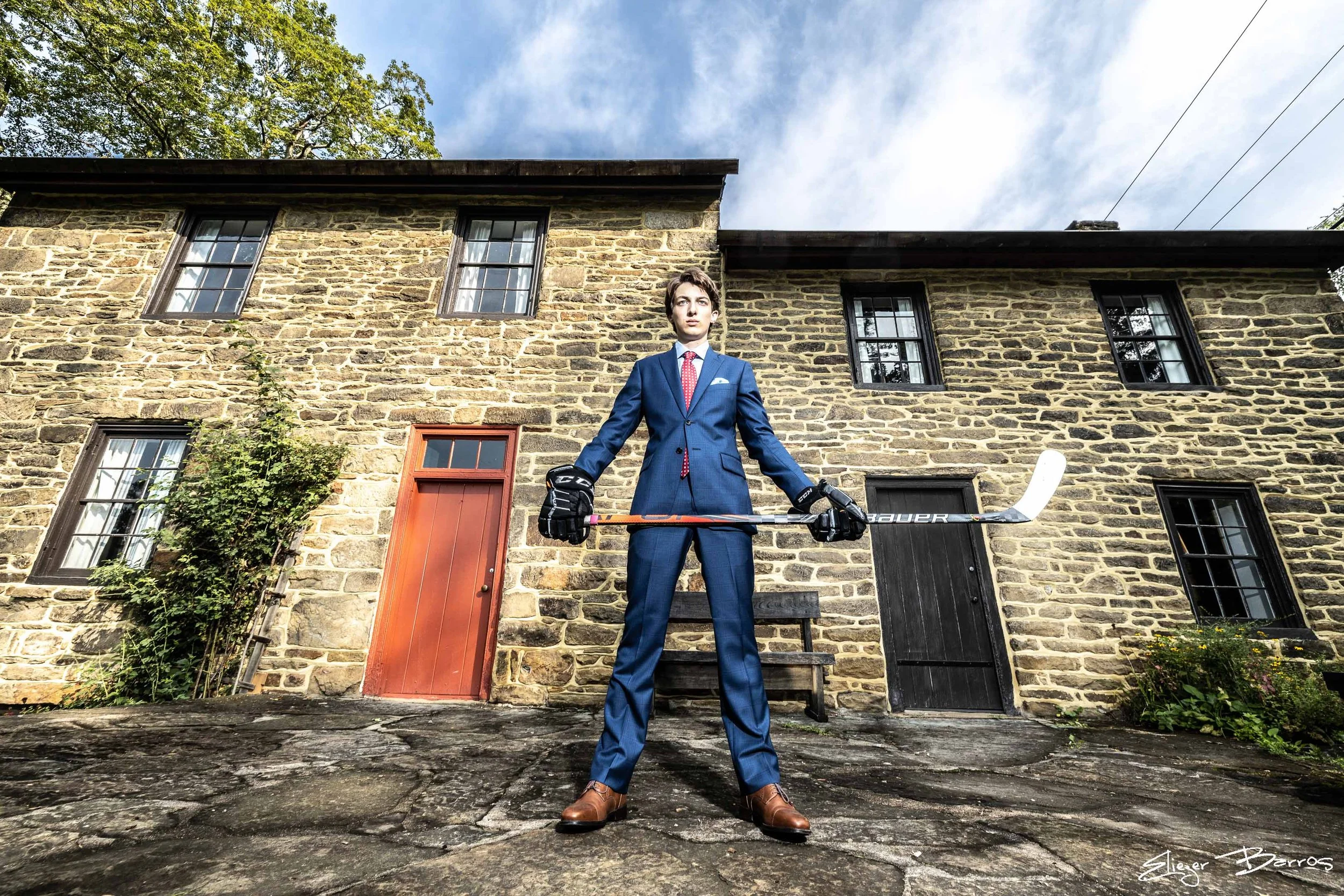 A young man dressed in a blue business suit holding a hockey stick outdoors in front of a stone building with black and red doors and multiple windows.