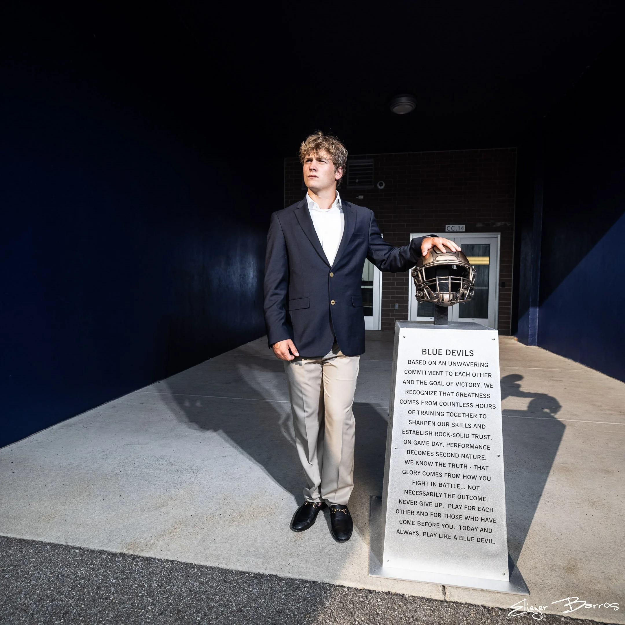 A young man in a formal suit holding a football stands next to an informational sign outside a building. The sign discusses the importance of commitment and teamwork, using the metaphor of the 'Blue Devils' and playing like a blue devil.