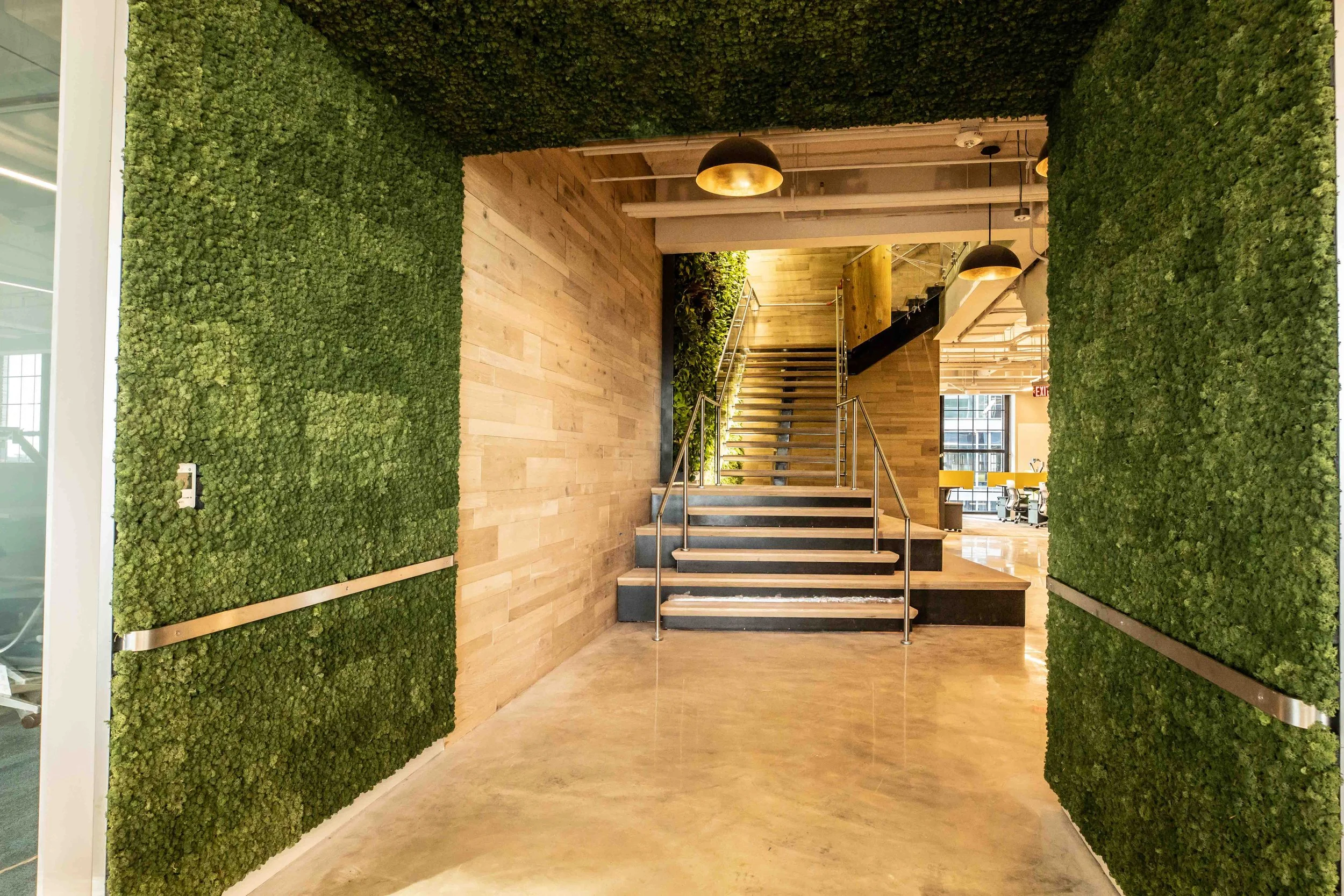 Interior of a modern office space with green walls made of moss or plants on both sides, wooden flooring, and a staircase with stainless steel handrails leading to an upper level, illuminated by round black and gold pendant lights, with large windows