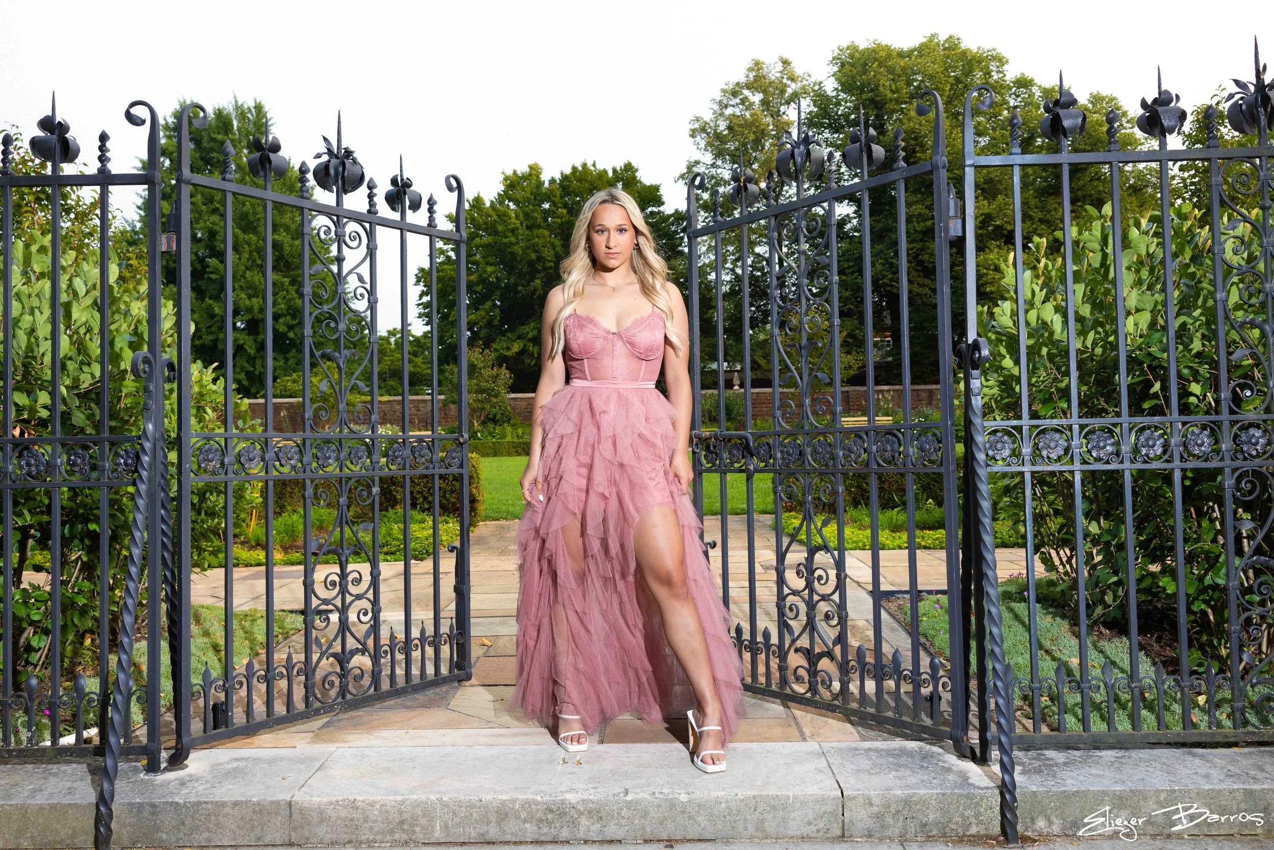 A young woman in a flowing pink dress stands in front of an ornate black iron gate in a garden setting, with green trees and bushes in the background.