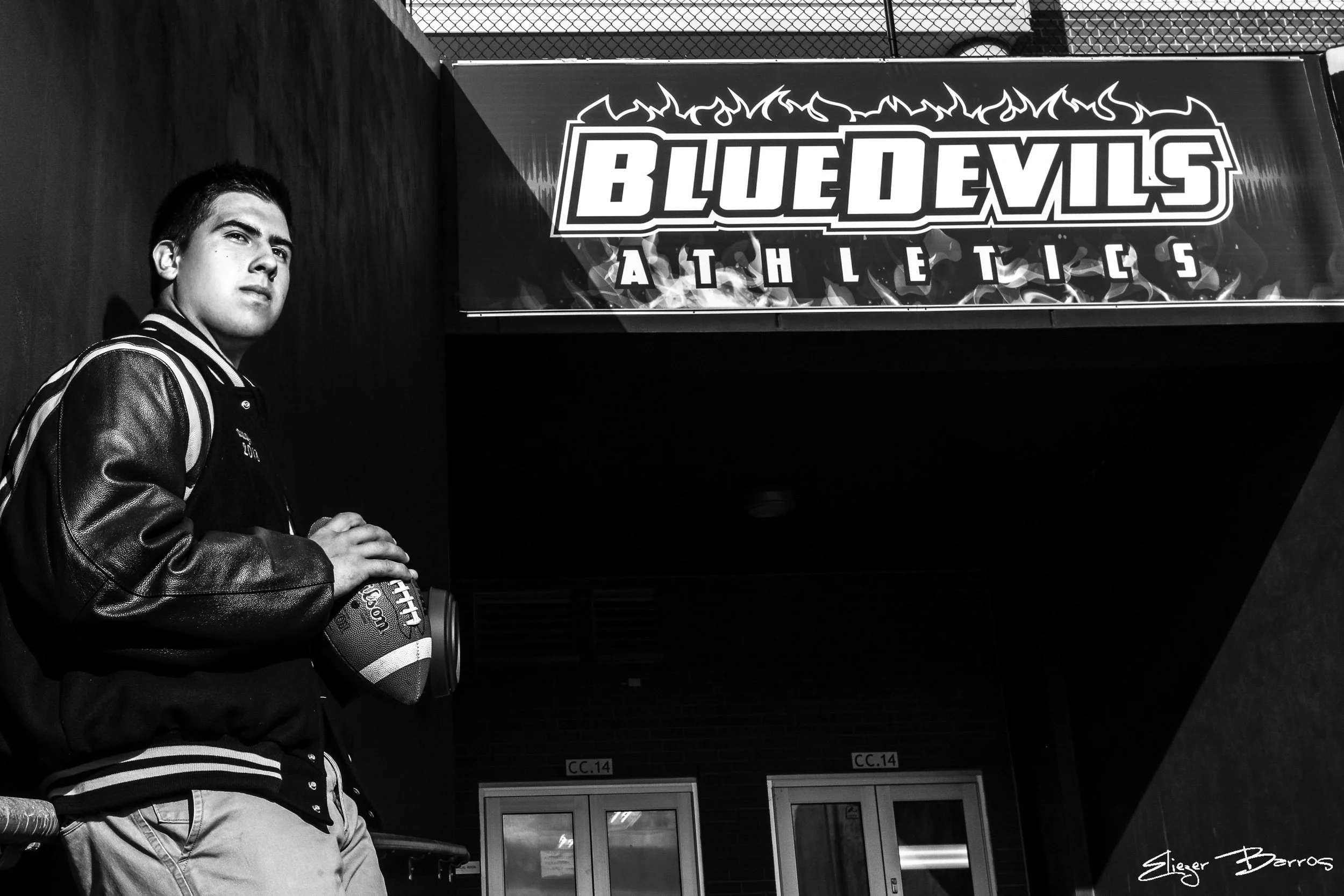 A young male athlete holding a football, wearing a varsity jacket, sitting near a sign that reads 'Blue Devils Athletics' with flames design, in an indoor sports facility.