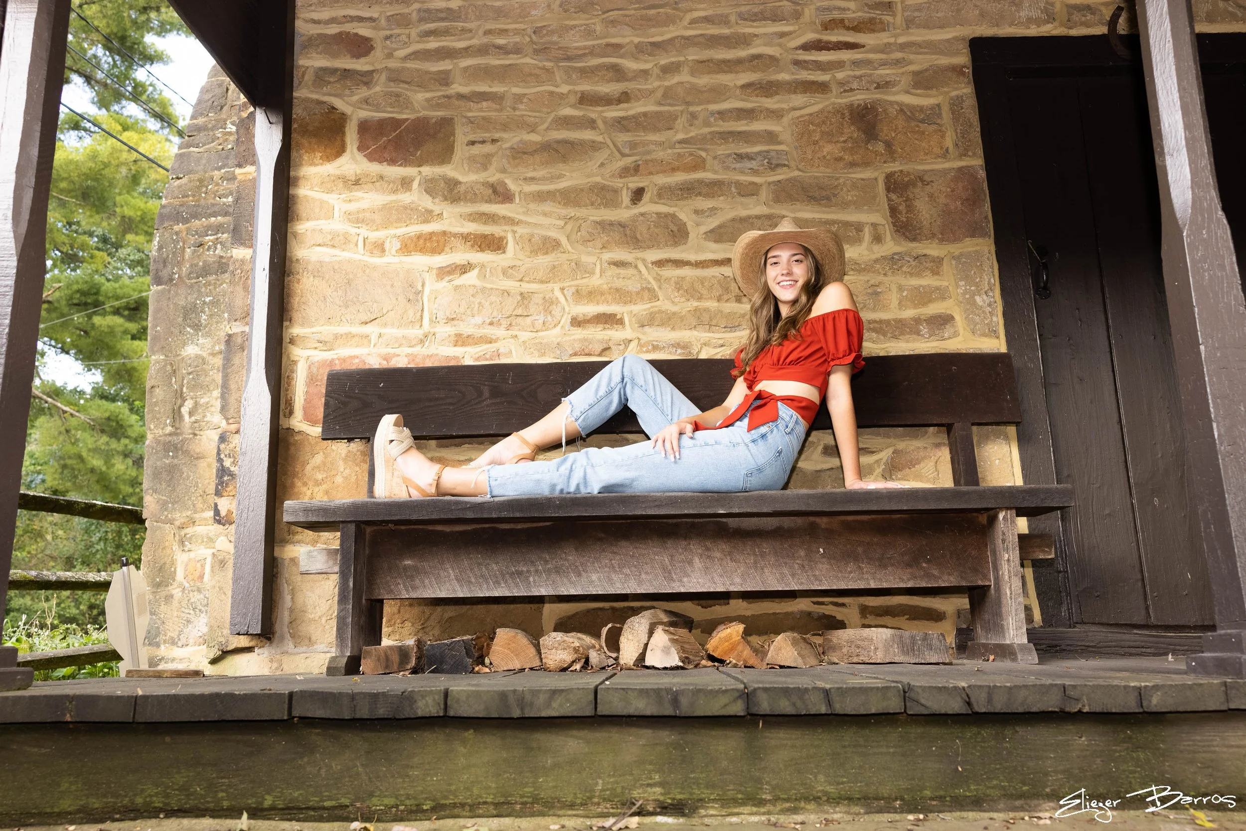 Young woman with long hair, wearing a red off-shoulder top, light blue jeans, tan shoes, and a wide-brimmed hat, sitting on a wooden bench on a rustic porch made of stone and wood, smiling at the camera.