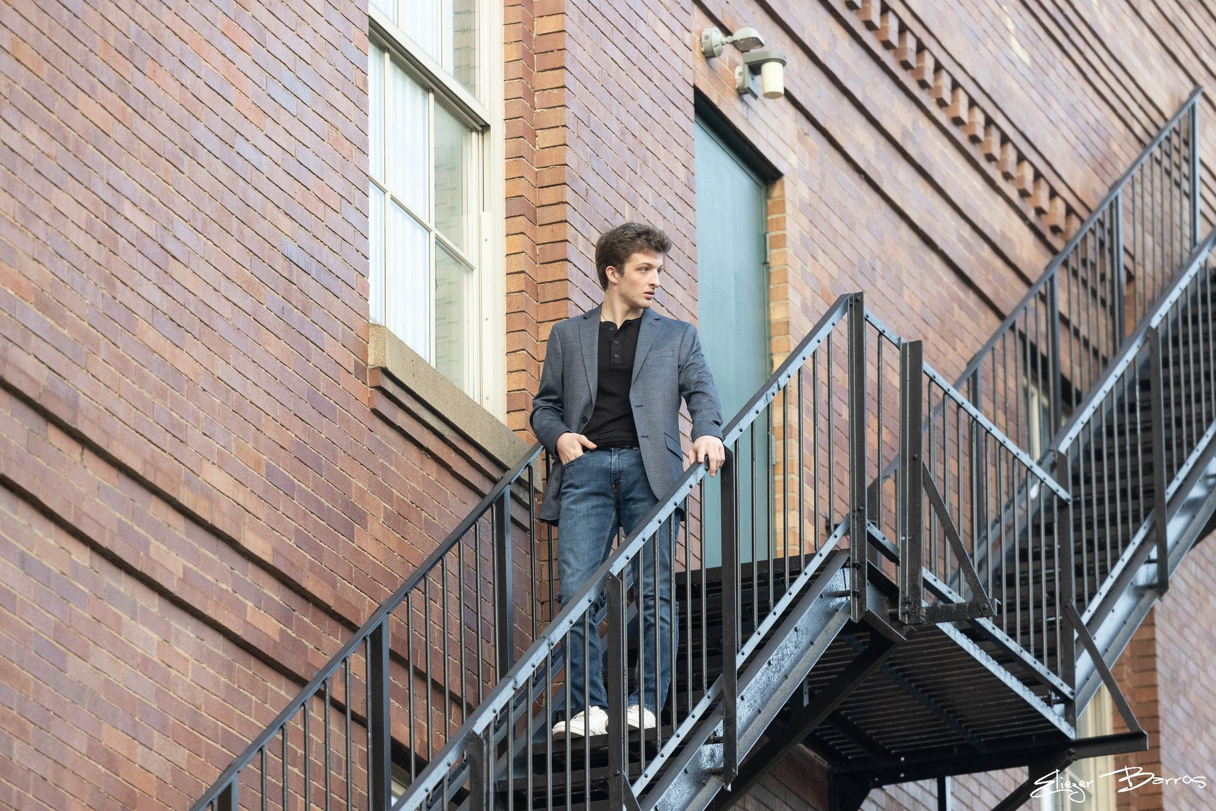 A young man in a gray blazer and jeans standing on an outdoor metal staircase next to a brick building with a window.