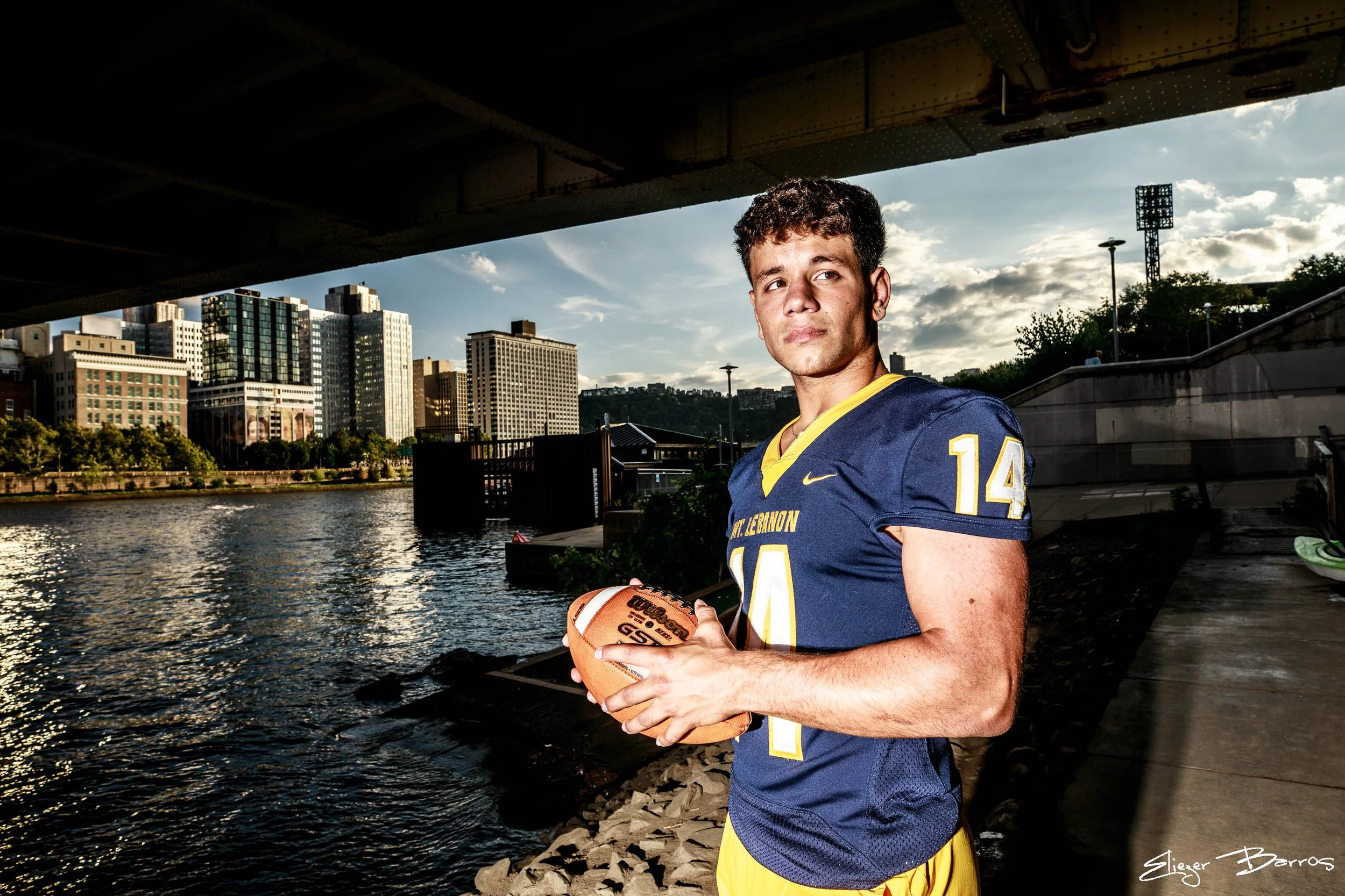 A young man in a football uniform holding a football by a river under a bridge in an urban setting.