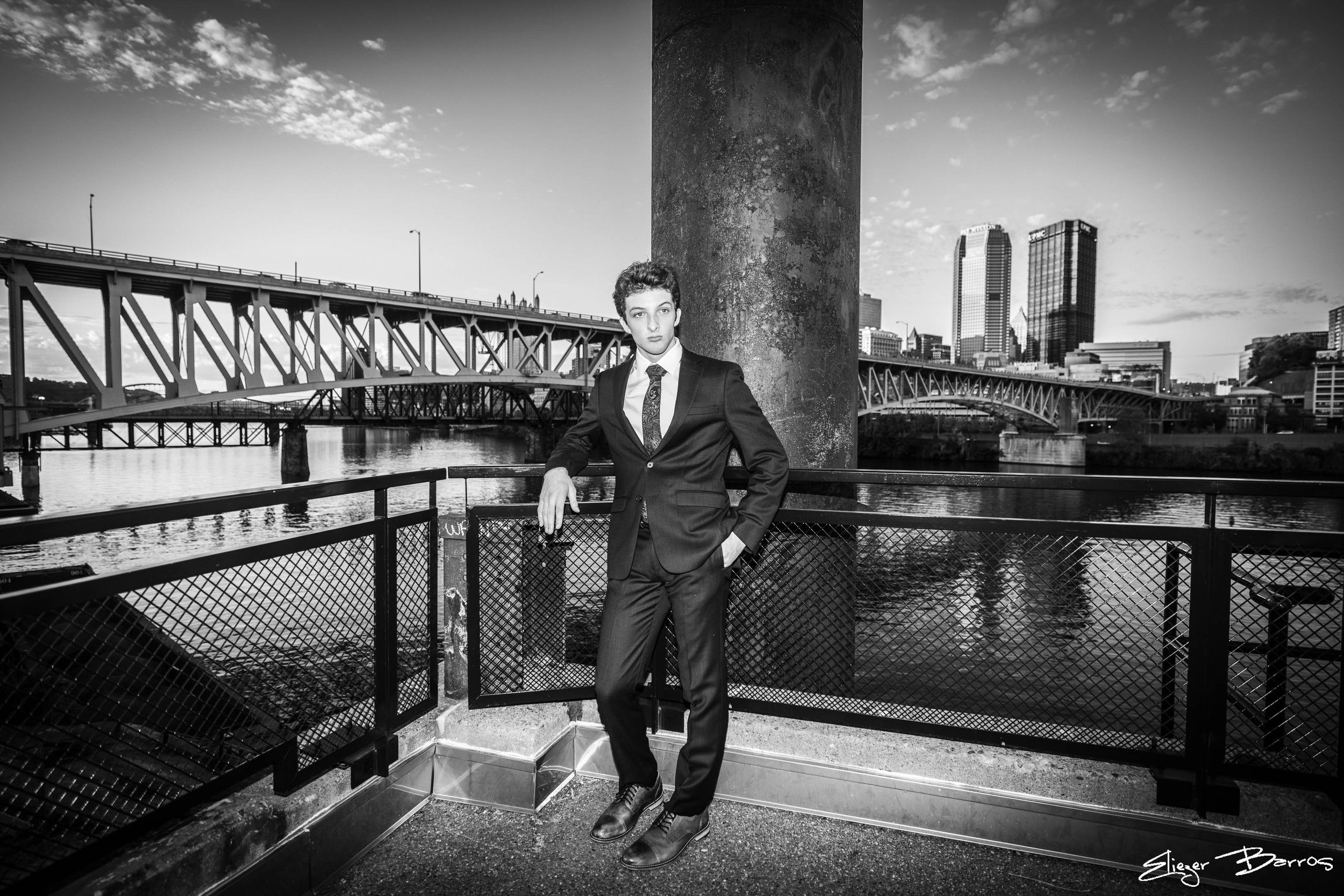 Black and white photo of a young man in a suit standing on a balcony by a river with a city skyline and bridge in the background.