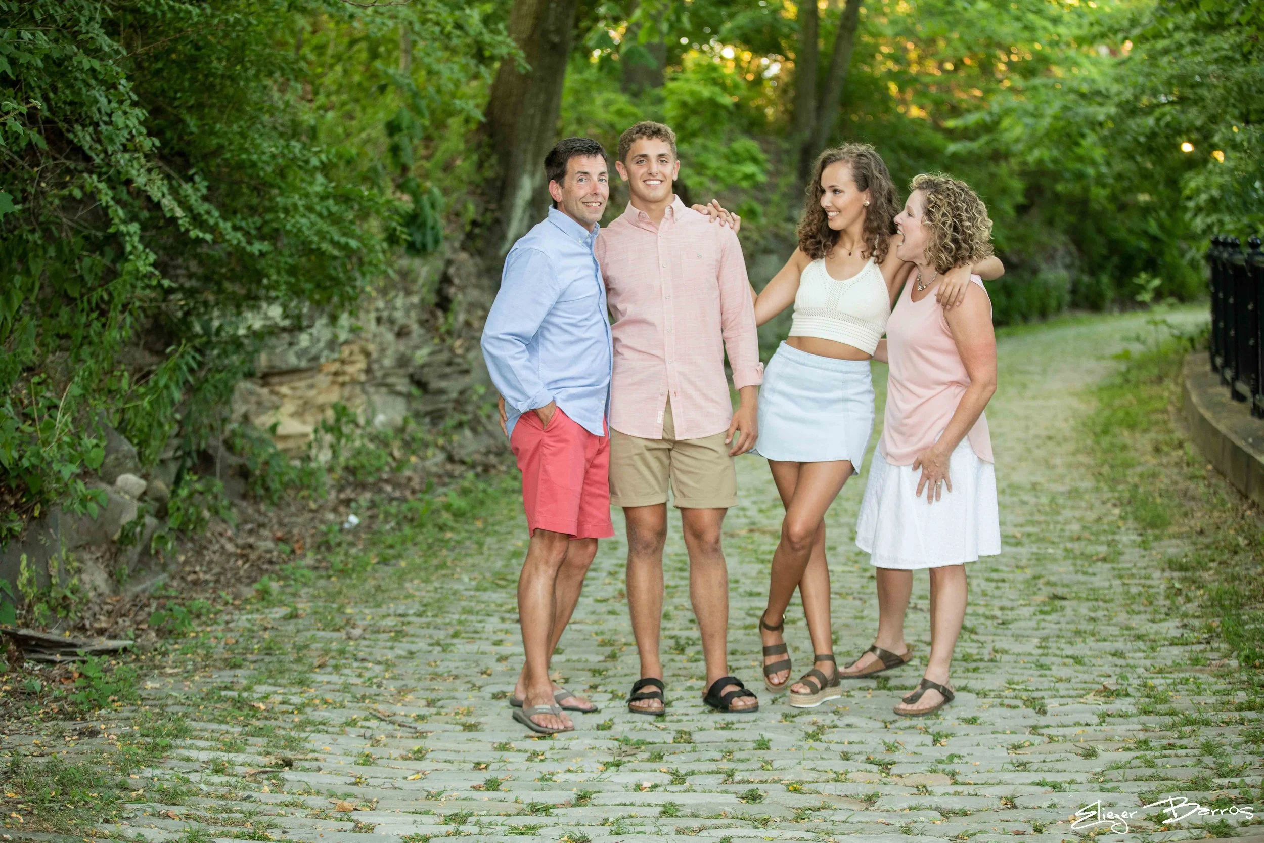 Four people standing on a cobblestone path surrounded by green trees, smiling and enjoying each other's company outdoors.