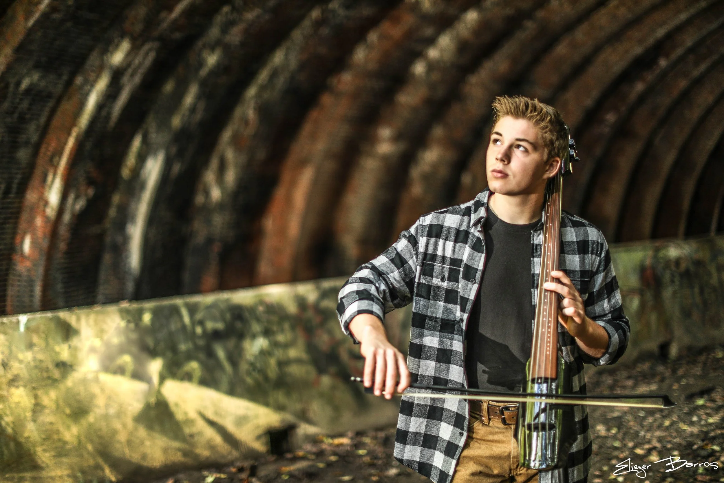 A young man with blonde hair wearing a black and white plaid shirt playing a double bass in a tunnel with brick walls.