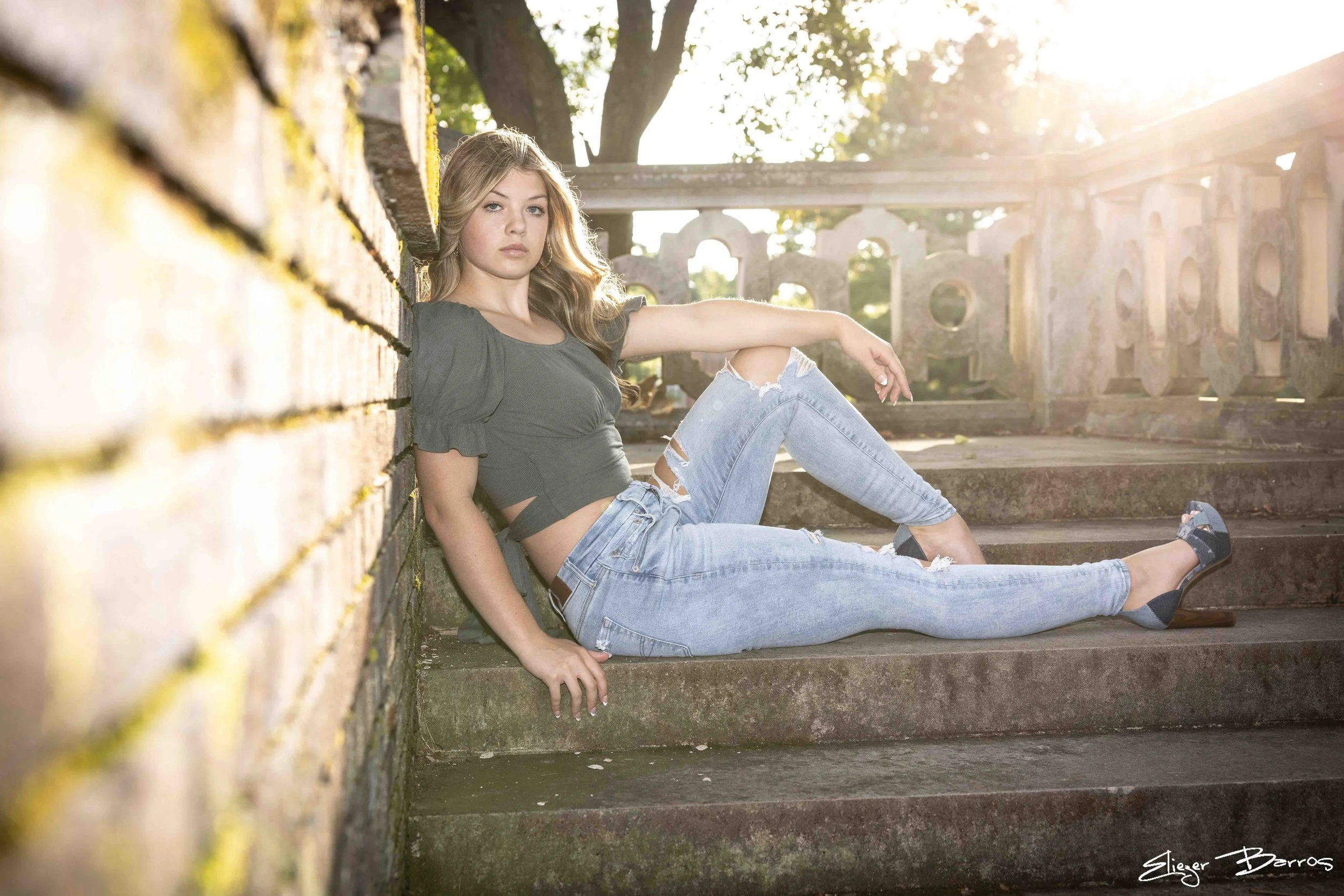 Young woman with wavy blonde hair sitting on stone steps outdoors, leaning against a brick wall. She is wearing a black top and ripped jeans, with one arm extended on a step and the other resting on the ground. Bright sunlight filters through trees i