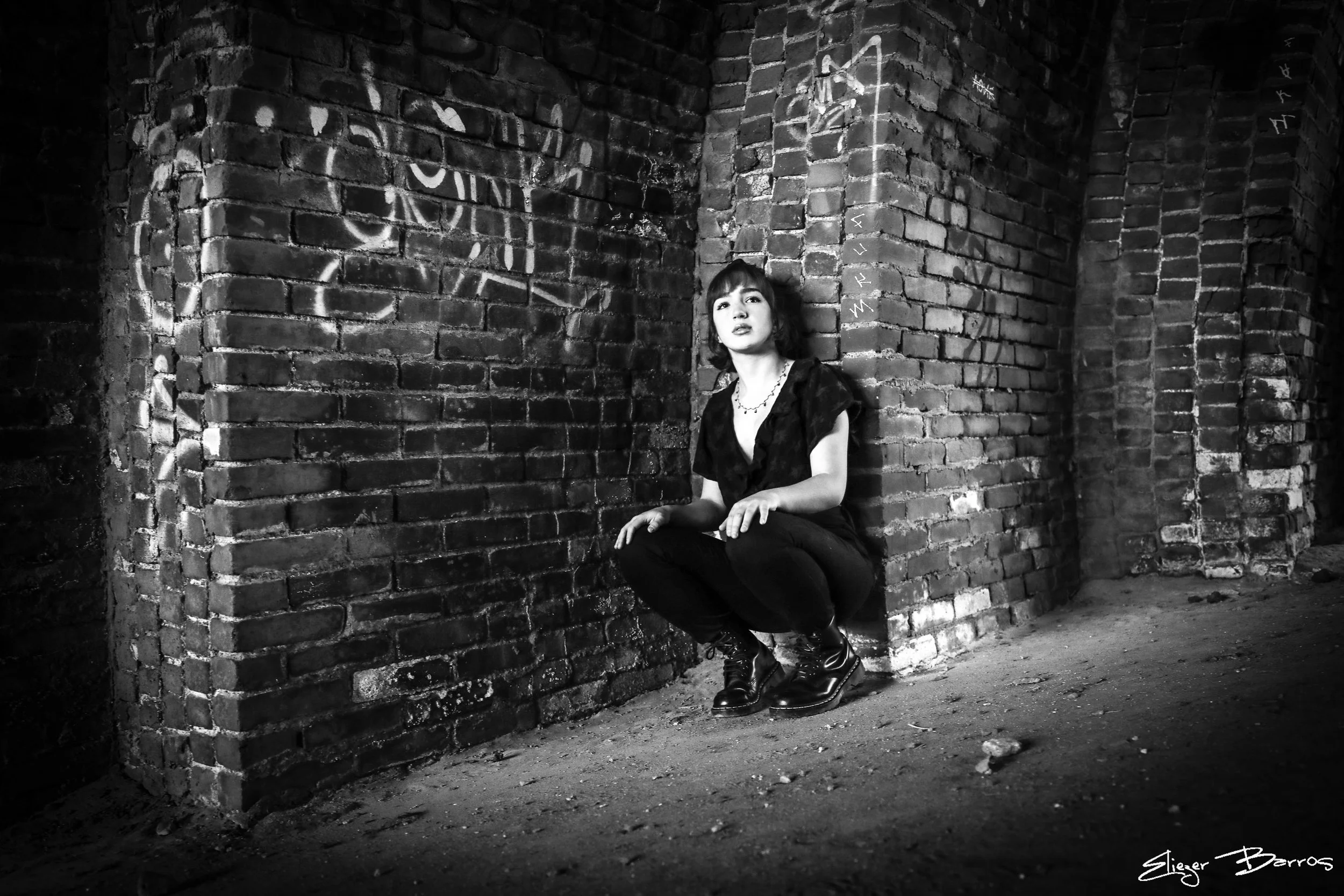 A black and white photo of a woman with dark hair and bangs, wearing a dark top and pants, sitting on her heels with her hands resting on her knees, against a brick corner of an alley or room. The brick wall behind her has graffiti. The ground is rou