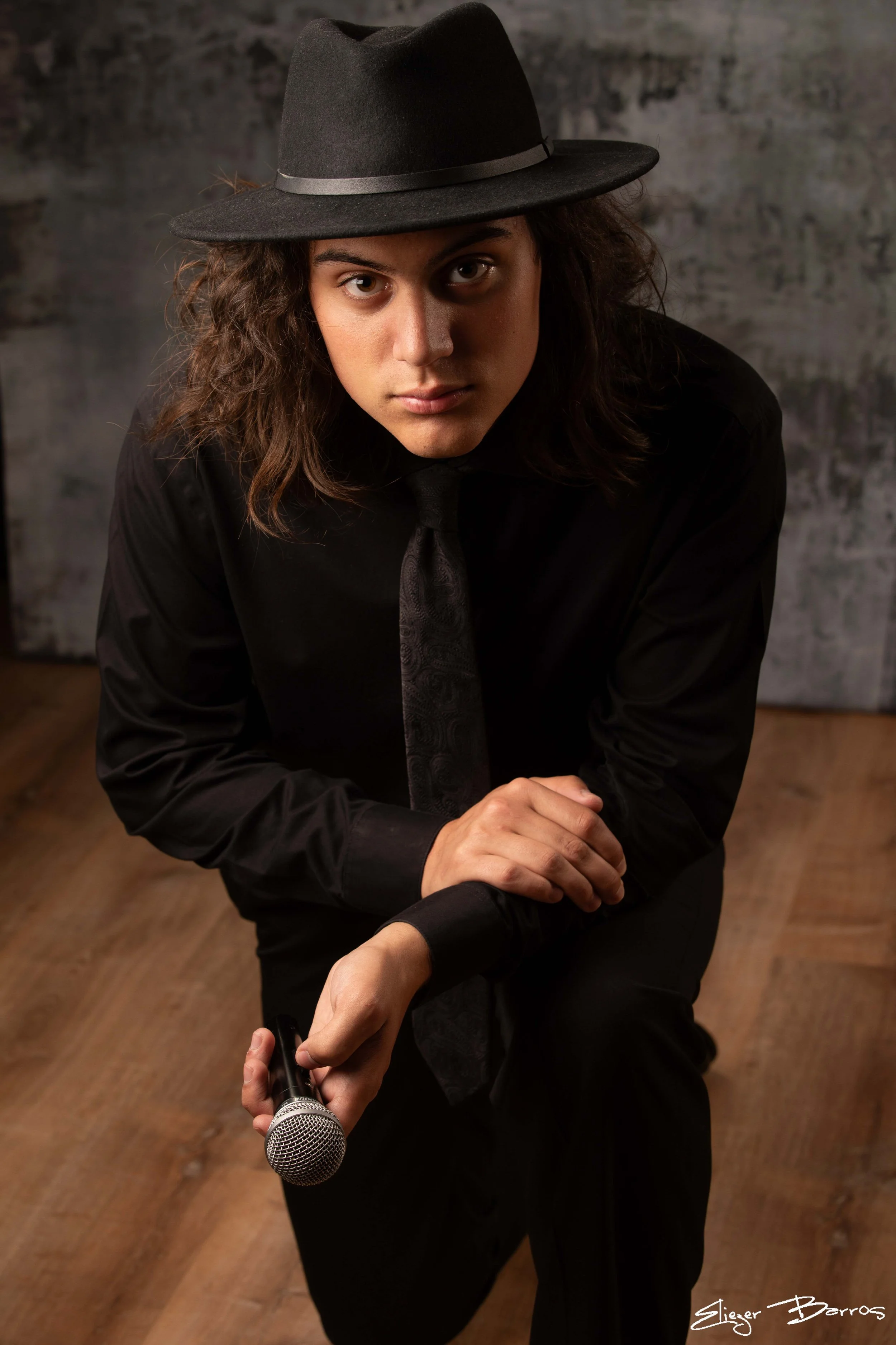 A person wearing a black fedora hat, black shirt, and dark tie, holding a microphone, kneeling on a wooden floor, with a dark textured background.