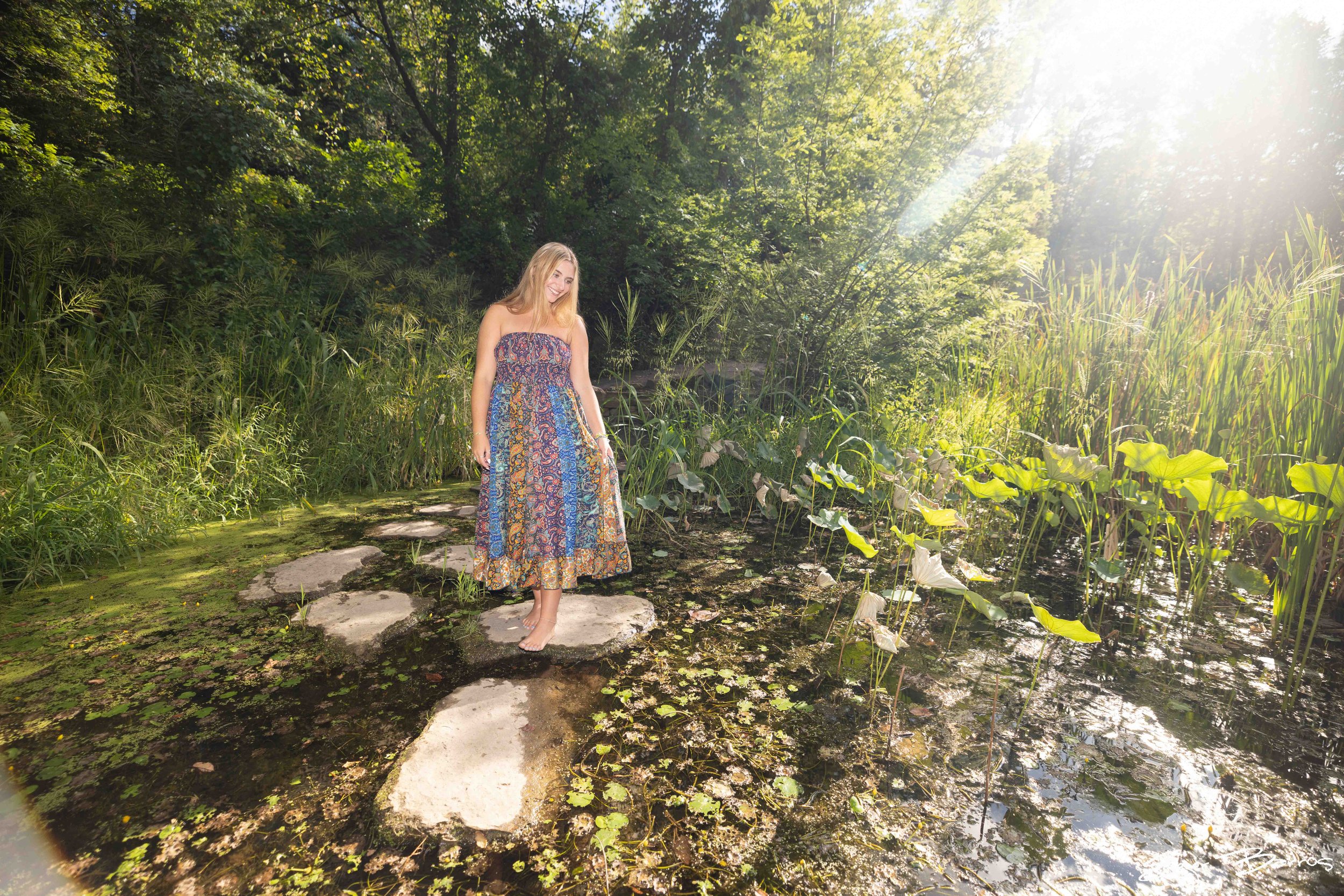 A woman in a colorful strapless dress walking barefoot on a stone path in a lush green wetland with sunlight filtering through trees.