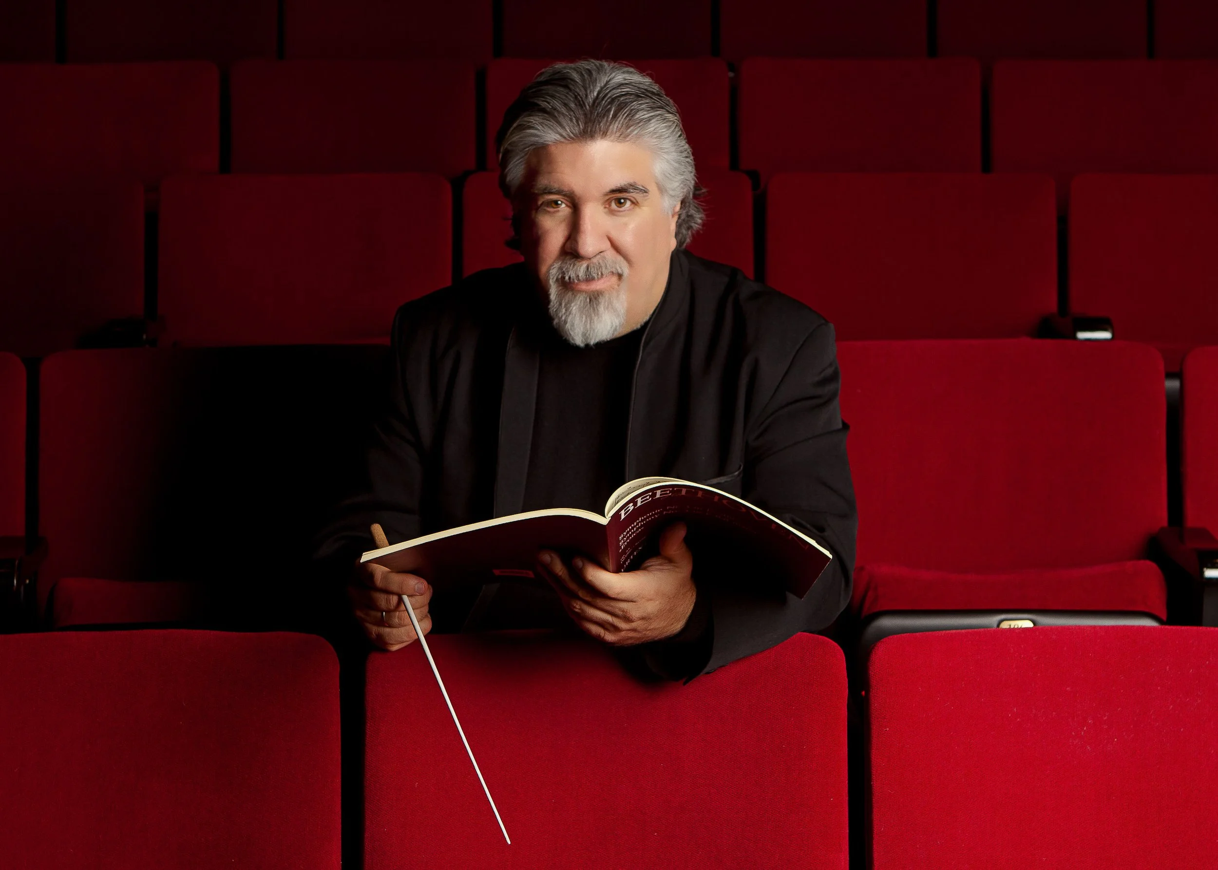 A middle-aged man with gray hair and a beard sitting in red theater seats, holding a book and a pen, with a focused expression.