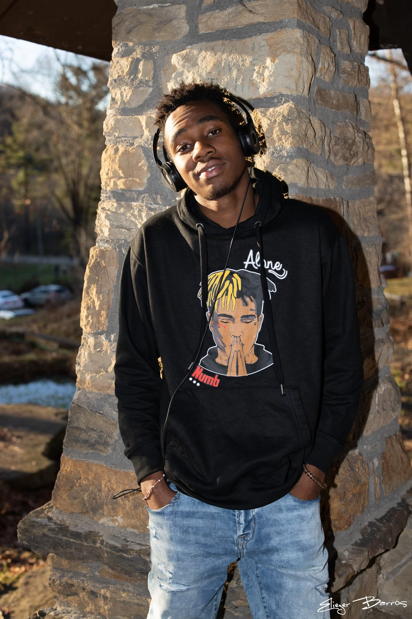 A young man with headphones around his neck, wearing a black hoodie with a cartoon face and the word 'Numb' on it, and ripped light blue jeans, standing outdoors against a stone pillar with trees and parked cars in the background.