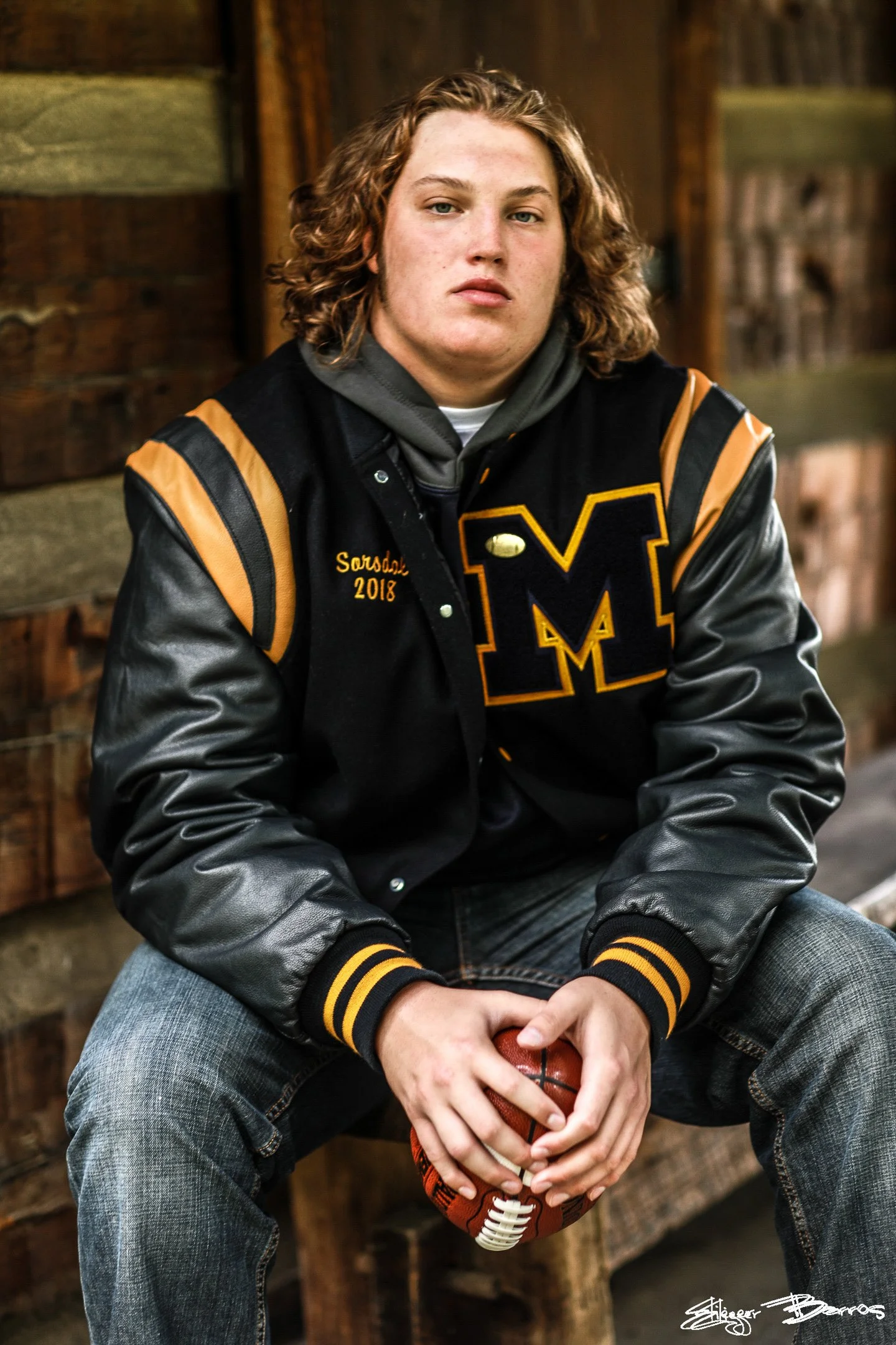 Senior Pictures Pittsburgh at Oliver Miller Homestead. Young man with curly hair in a black and gold varsity jacket sitting and holding a football in front of the historic stone building for nostalgic athletic senior portraits.