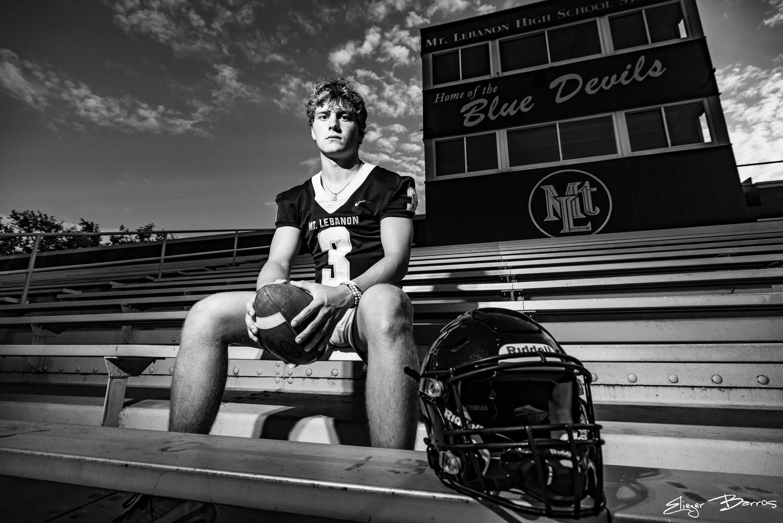 A young male football player sitting on bleachers at a football field, wearing a uniform with the name 'Mt. Lebanon' and the number 3. He is holding a football, with a helmet and a backpack with a helmet nearby. The photo is in black and white, with 