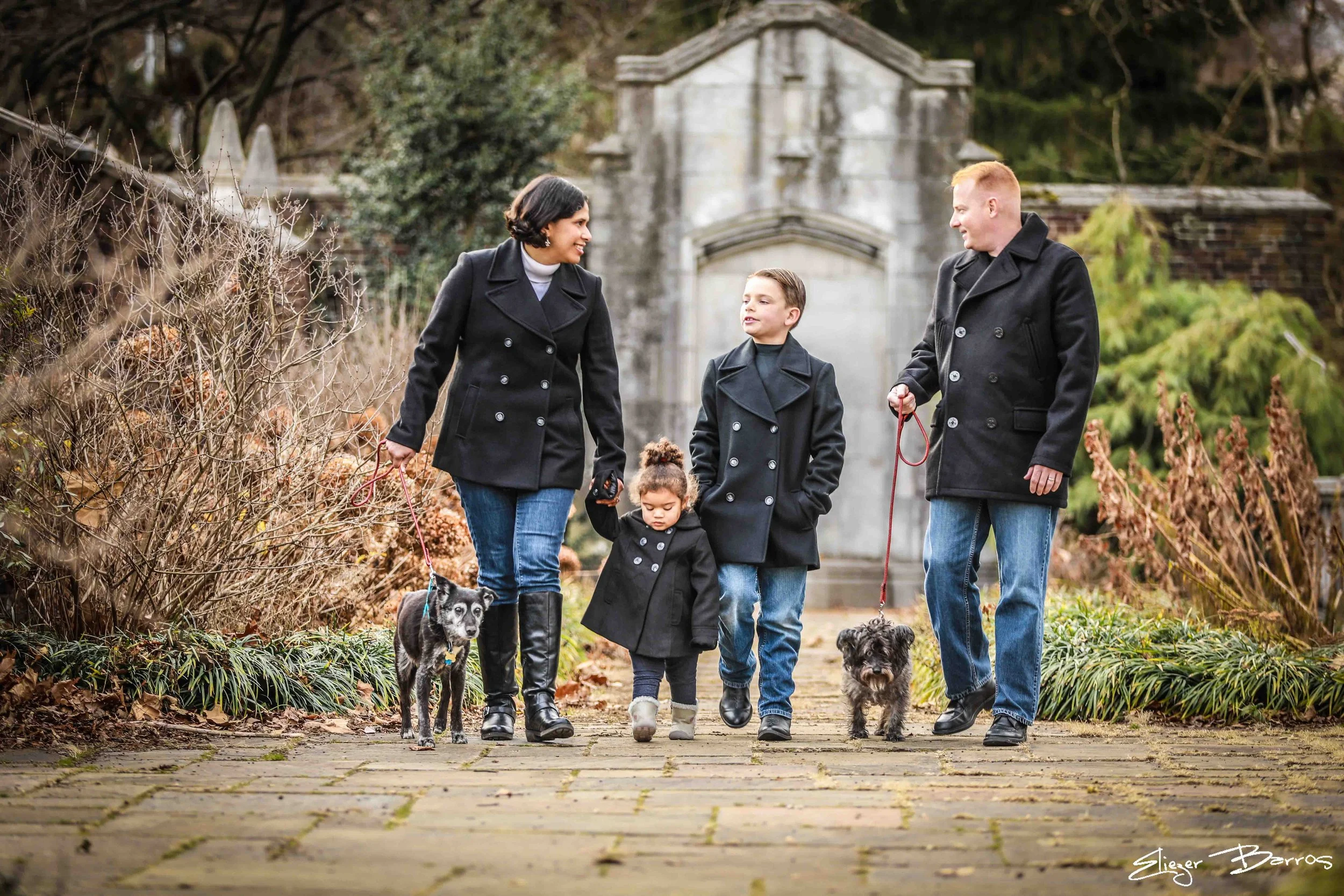 A family of four walking with two dogs on a paved pathway through a park or garden. The family includes a woman, a man, and two children. All are dressed in black coats, and the children are holding the dogs' leashes. The background features autumnal