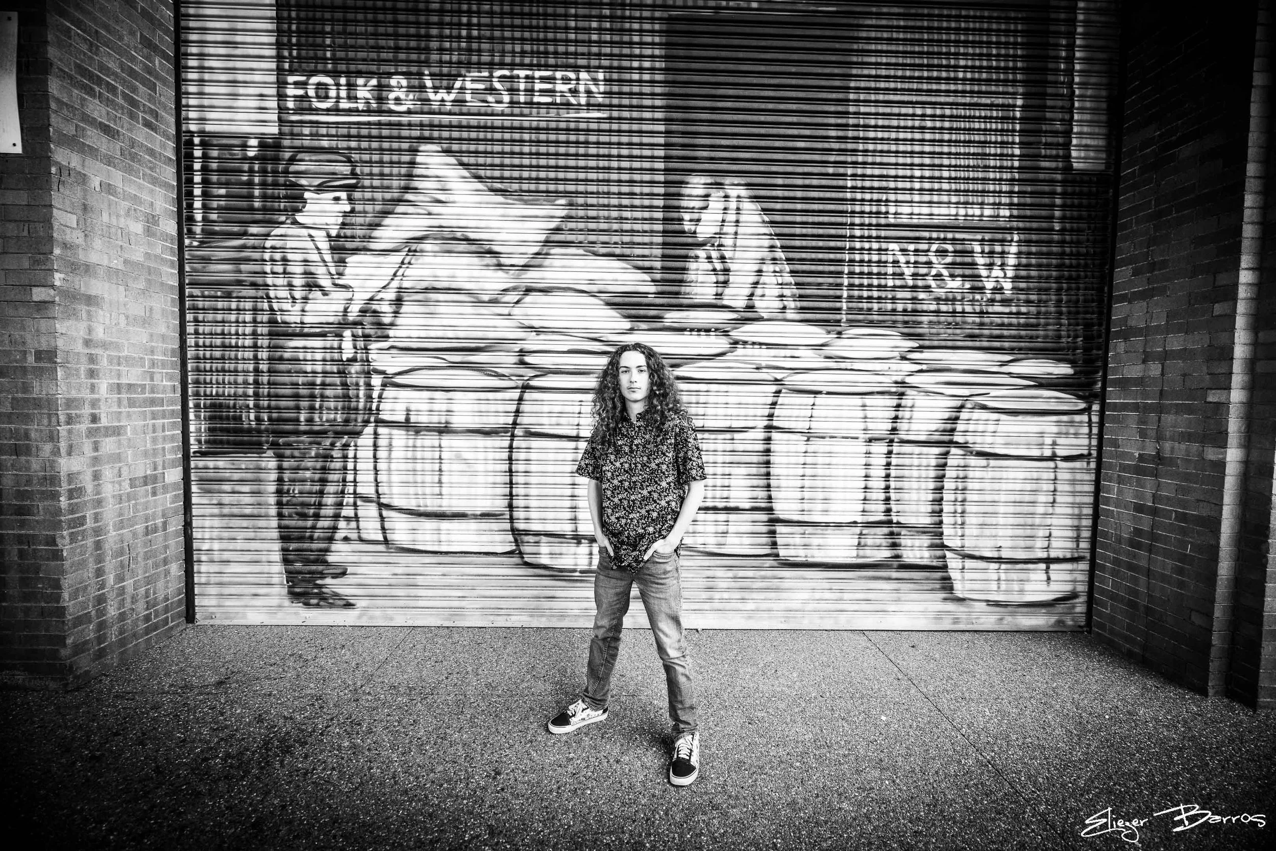 A woman with curly hair wearing a floral shirt, jeans, and sneakers standing in front of a mural graffiti art of a person playing a guitar at night.
